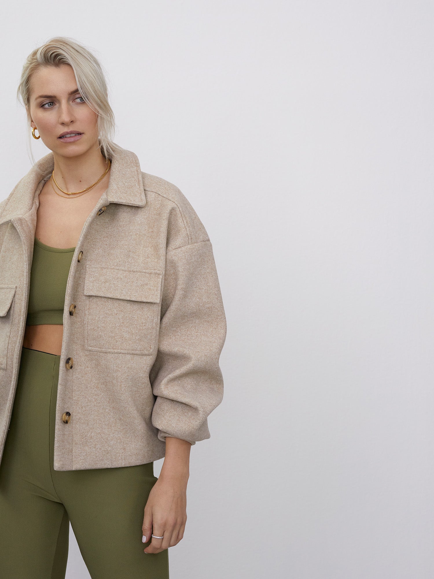 Eine blonde Person trägt die Jacke „Eva“ von LeGer by Lena Gercke in Beige über einem olivgrünen Oberteil und einer Hose, steht mit goldenen Ohrringen und einer Halskette vor einem schlichten weißen Hintergrund und blickt zur Seite.