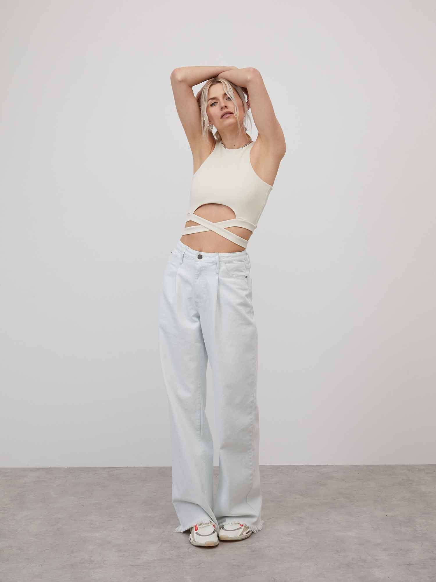 Eine Person modelt in einem minimalistischen Studio und trägt das ärmellose, cremefarbene Top „Shelly“ von LeGer by Lena Gercke mit kurzem Schnitt und Cut-Out-Design. Sie kombiniert es mit hellblauen Jeans mit weitem Bein und streckt die Arme über den Kopf vor einem schlichten hellgrauen Hintergrund.