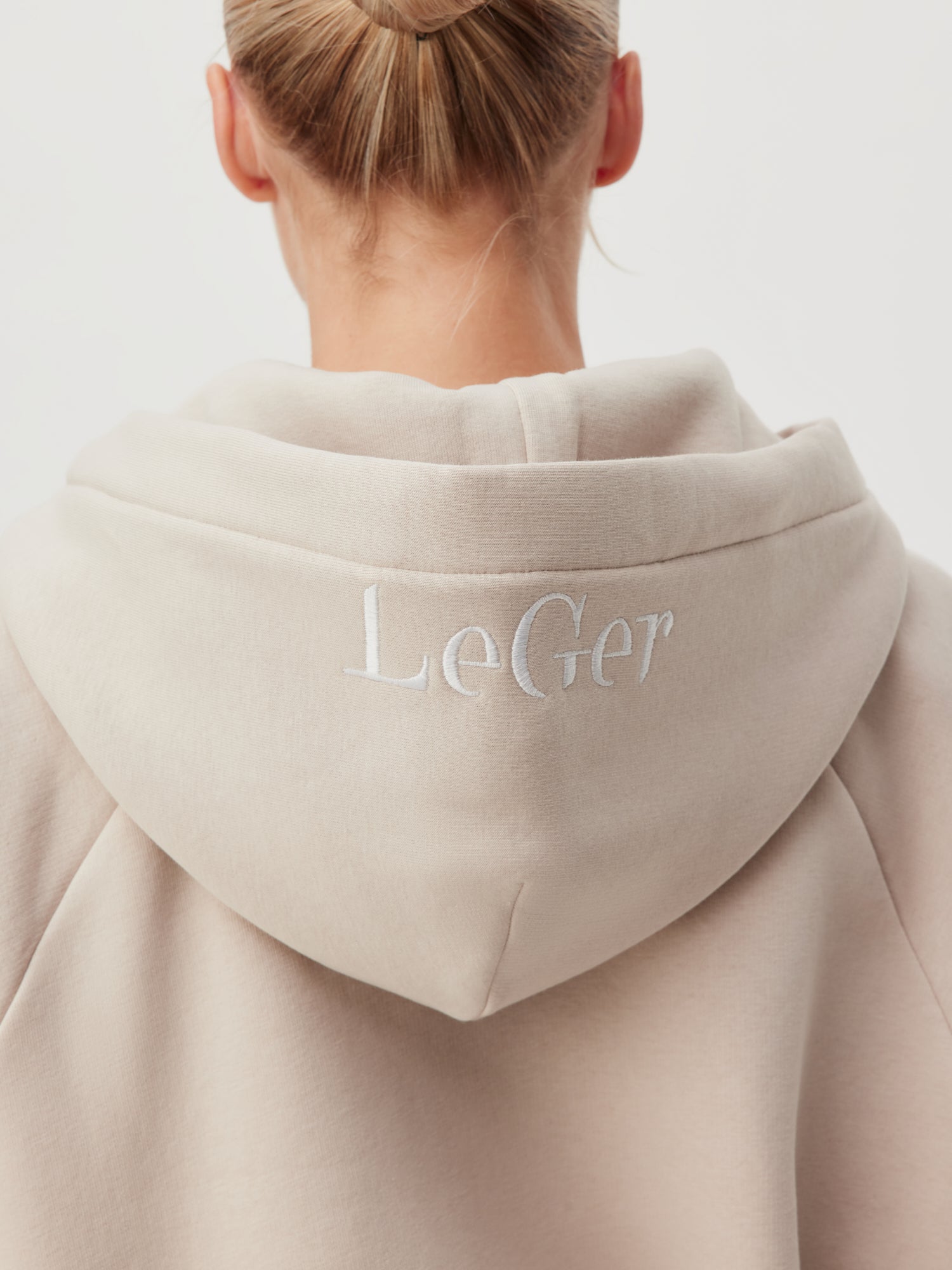 Auf der Rückseite ist eine Person mit blonden Haaren zu sehen, die ein hellbeiges Kapuzensweatshirt namens „Hayley“ von LeGer by Lena Gercke trägt. Auf der Rückseite der Kapuze ist in Weiß der Schriftzug „LeGer“ aufgestickt, was das stylische Langarm-Design hervorhebt.