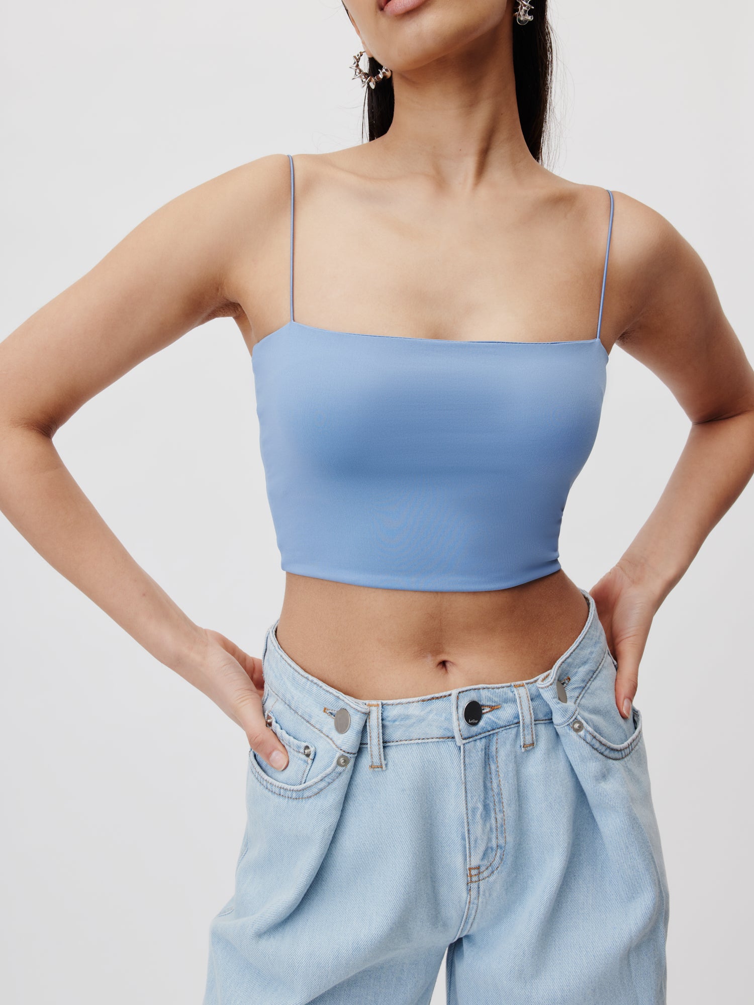 Eine Person, die ein hellblaues Joelle-Top von LeGer by Lena Gercke, ein ärmelloses Crop-Top mit Spaghettiträgern und hoch taillierte, schmale hellblaue Jeans trägt, steht mit den Händen auf den Hüften vor einem schlichten weißen Hintergrund.