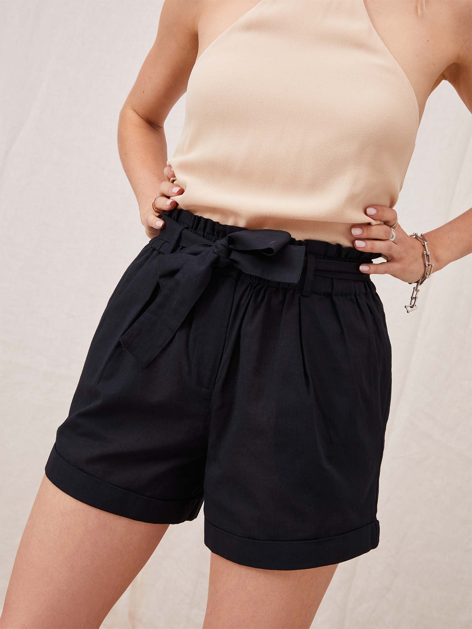 Eine Frau trägt ein beiges Neckholder-Top mit Yasmina-Shorts von LeGer by Lena Gercke, mit hoher Taille und gebundenem Gürtel in schwarzer Minilänge. Ihre Hände ruhen auf ihren Hüften, geschmückt mit einem Armband an ihrem rechten Handgelenk. Der Hintergrund ist hell und neutral.