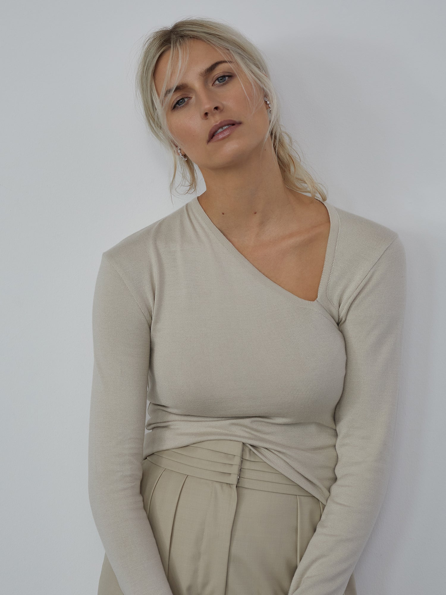 Eine Person mit langen blonden Haaren trägt das Helen Top von LeGer by Lena Gercke, ein beiges One-Shoulder-Design, das perfekt zu ihren hoch taillierten Hosen passt. Das Ensemble hat einen Passform Schmal-Look, während sie mit entspanntem Gesichtsausdruck vor einer schlichten weißen Wand steht.