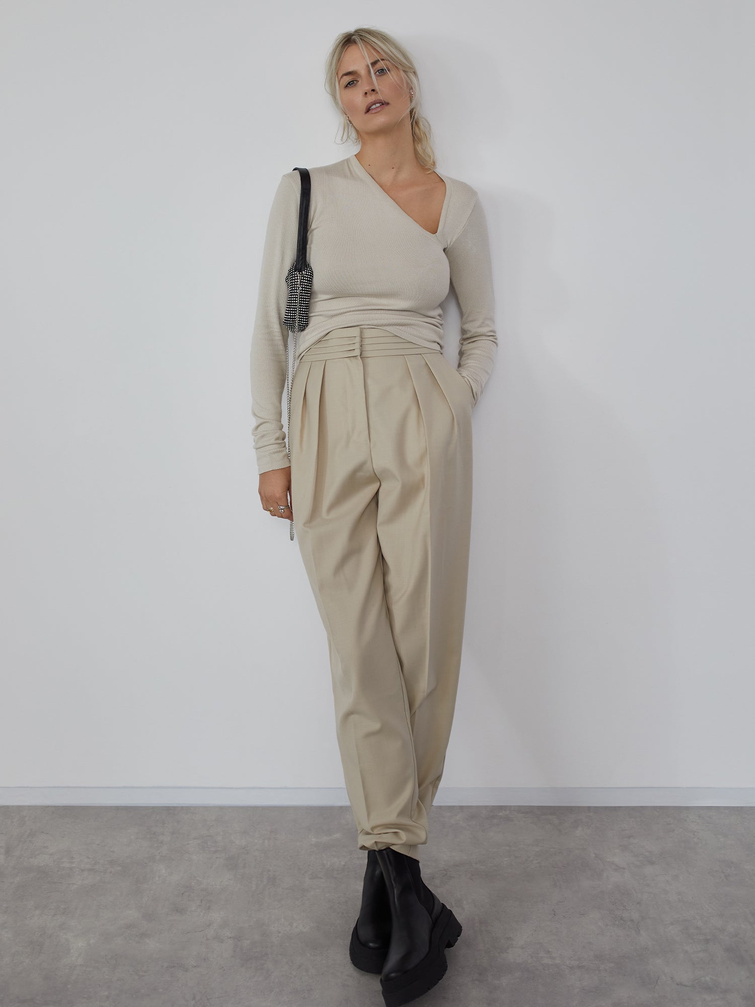 Eine Person posiert vor einer weißen Wand. Sie trägt das Helen Top von LeGer by Lena Gercke, ein beiges asymmetrisches Top mit Langarm, kombiniert mit hoch taillierten Bundfaltenhosen in Passform Schmal. Sie hat ihre Hände lässig in den Taschen verstaut, trägt schwarze Stiefel und eine elegante, lockere Hochsteckfrisur, die ihre Umhängetasche ergänzt.