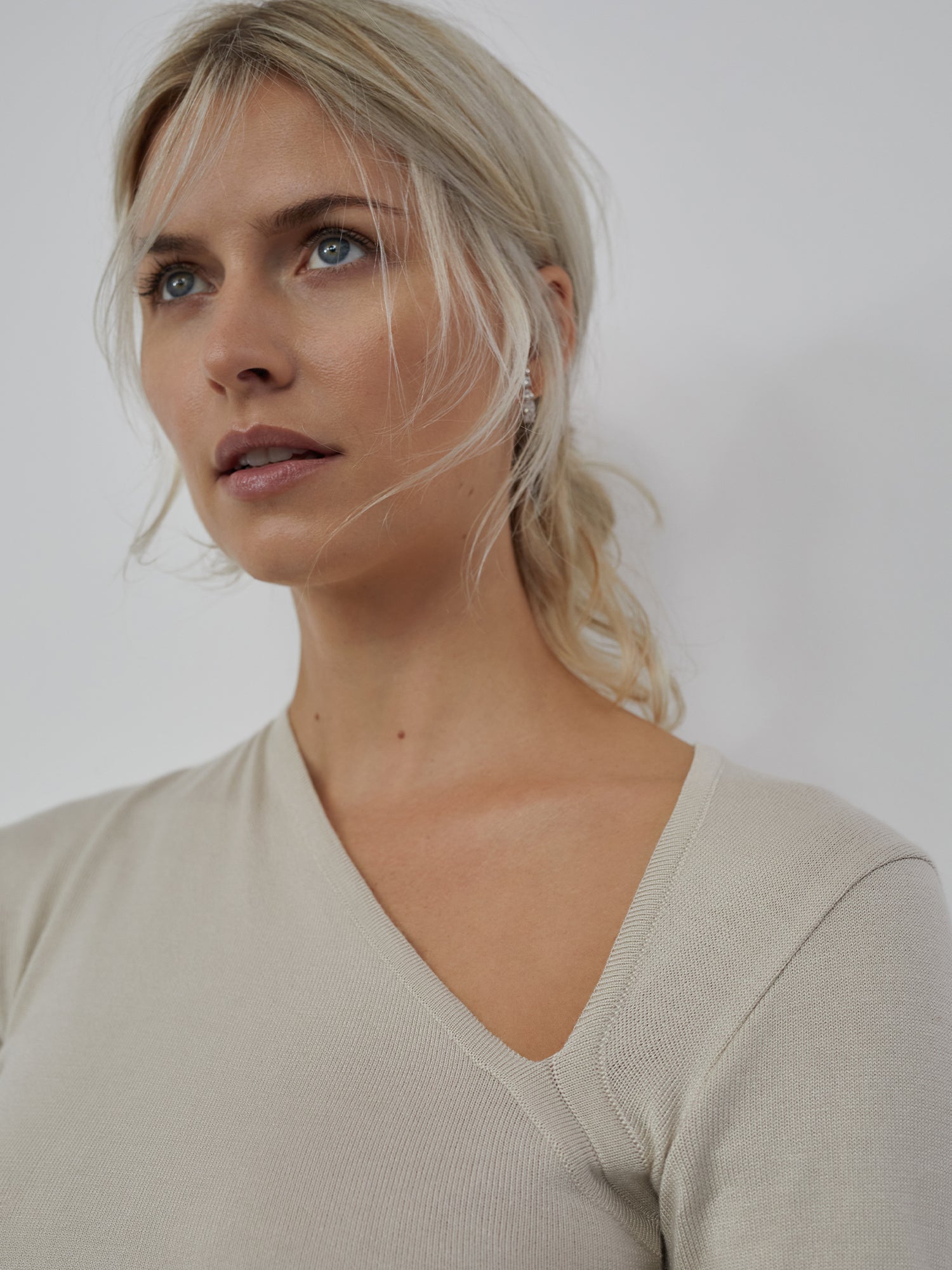 Eine Person mit zurückgebundenem blondem Haar trägt das Helen Top, ein beiges V-Ausschnitt-Design von LeGer by Lena Gercke, mit langen Ärmeln und schmaler Passform, und blickt vor einem einfarbigen Hintergrund leicht nach oben.