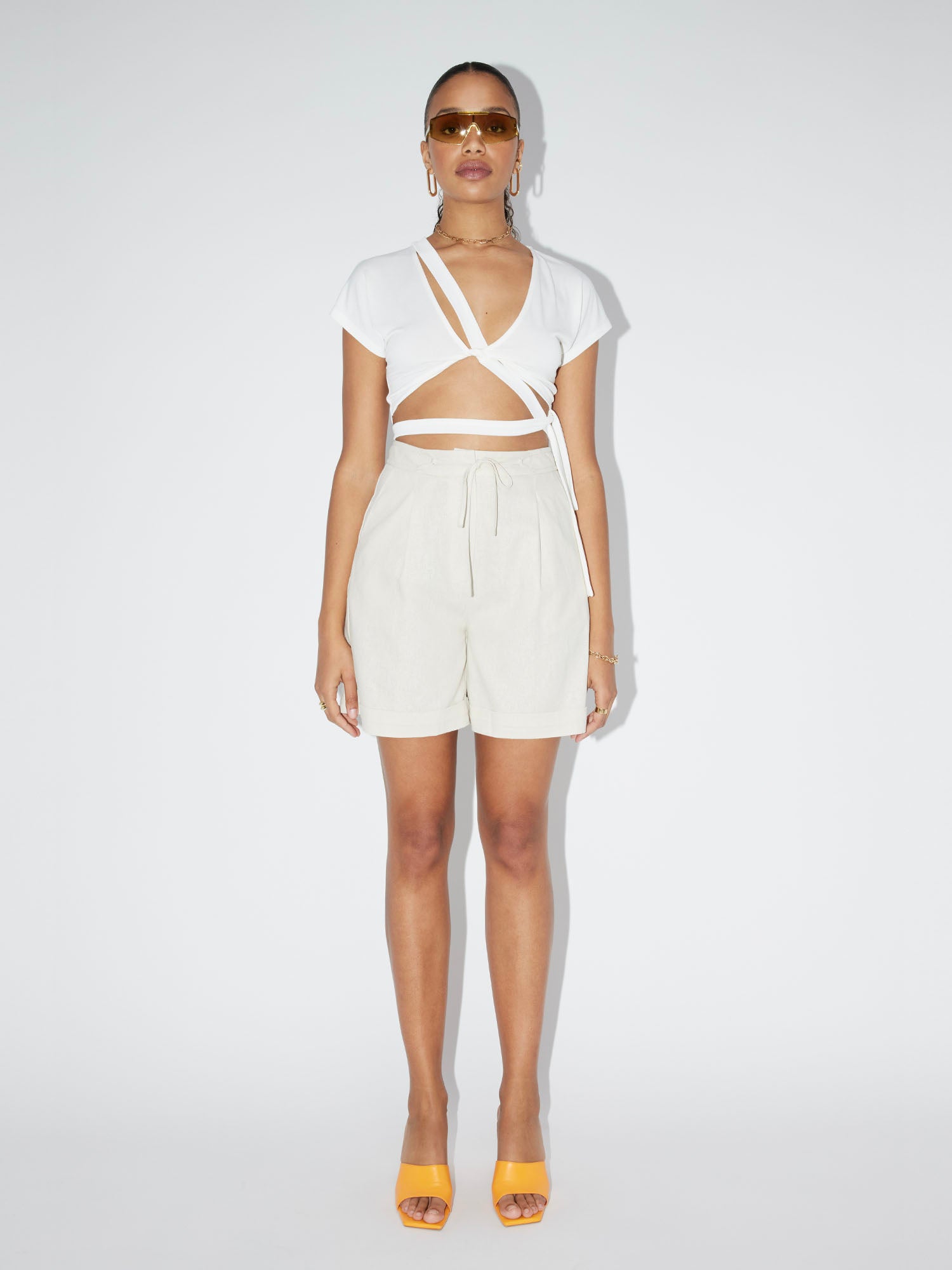 Eine Person posiert vor einem schlichten Hintergrund. Sie trägt eine Sonnenbrille, ein weißes, kurzärmliges Crop-Top mit Wickeldesign und perfekter Passform, helle, hoch taillierte Shorts von LeGer by Lena Gercke namens „Imen“ und orangefarbene Stöckelschuhe. Ihre Haare sind zurückgekämmt und sie tragen Ohrringe und eine Halskette als Accessoires.