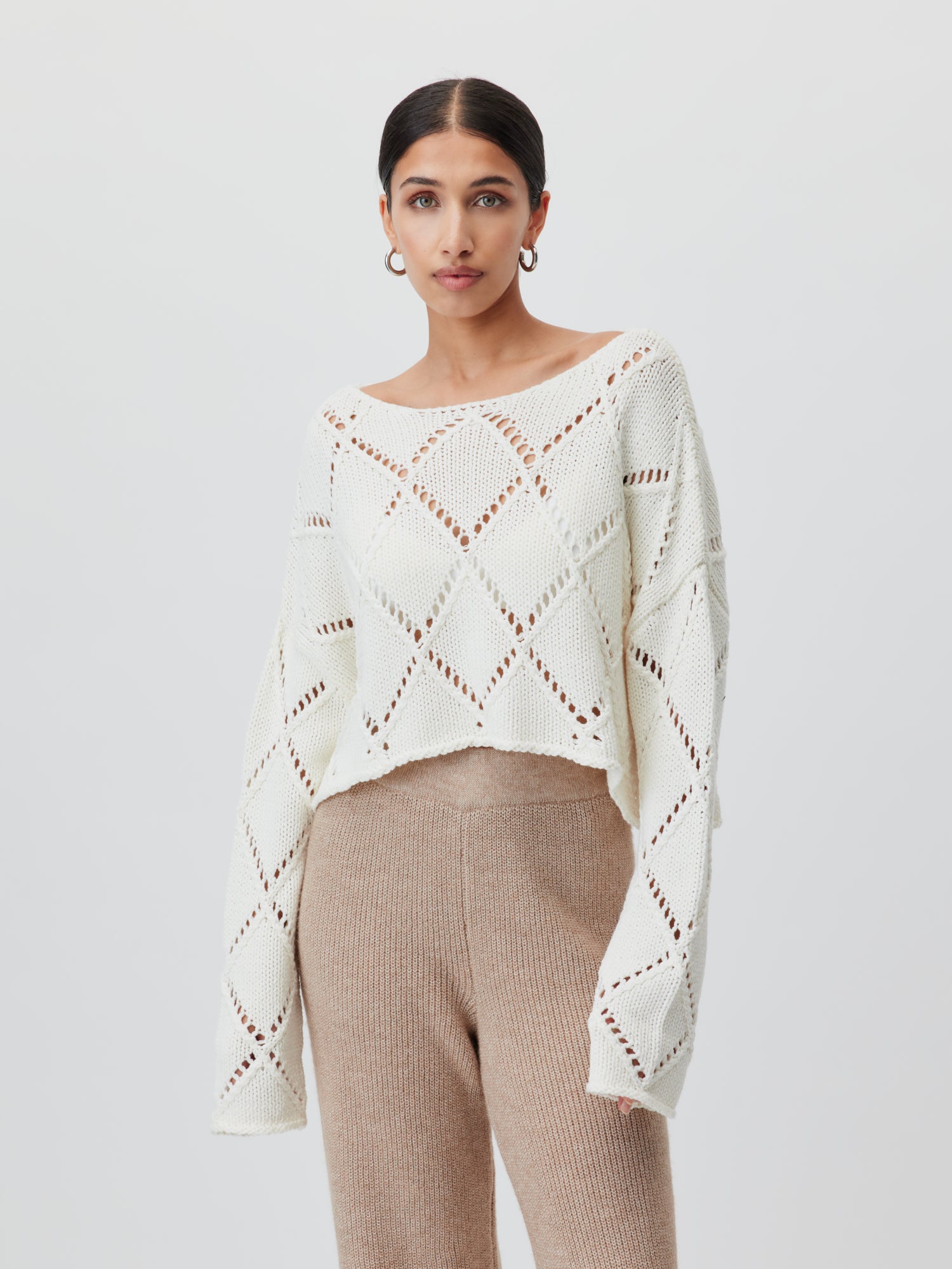 Eine Person mit dunklem Haar trägt den Briley Jumper von LeGer by Lena Gercke, einen weißen Strickpullover normaler Länge mit Rautenmuster, kombiniert mit hellbraunen Strickhosen. Die Passform des Pullovers ergänzt ihren Stil perfekt, da sie vor einem schlichten weißen Hintergrund stehen.