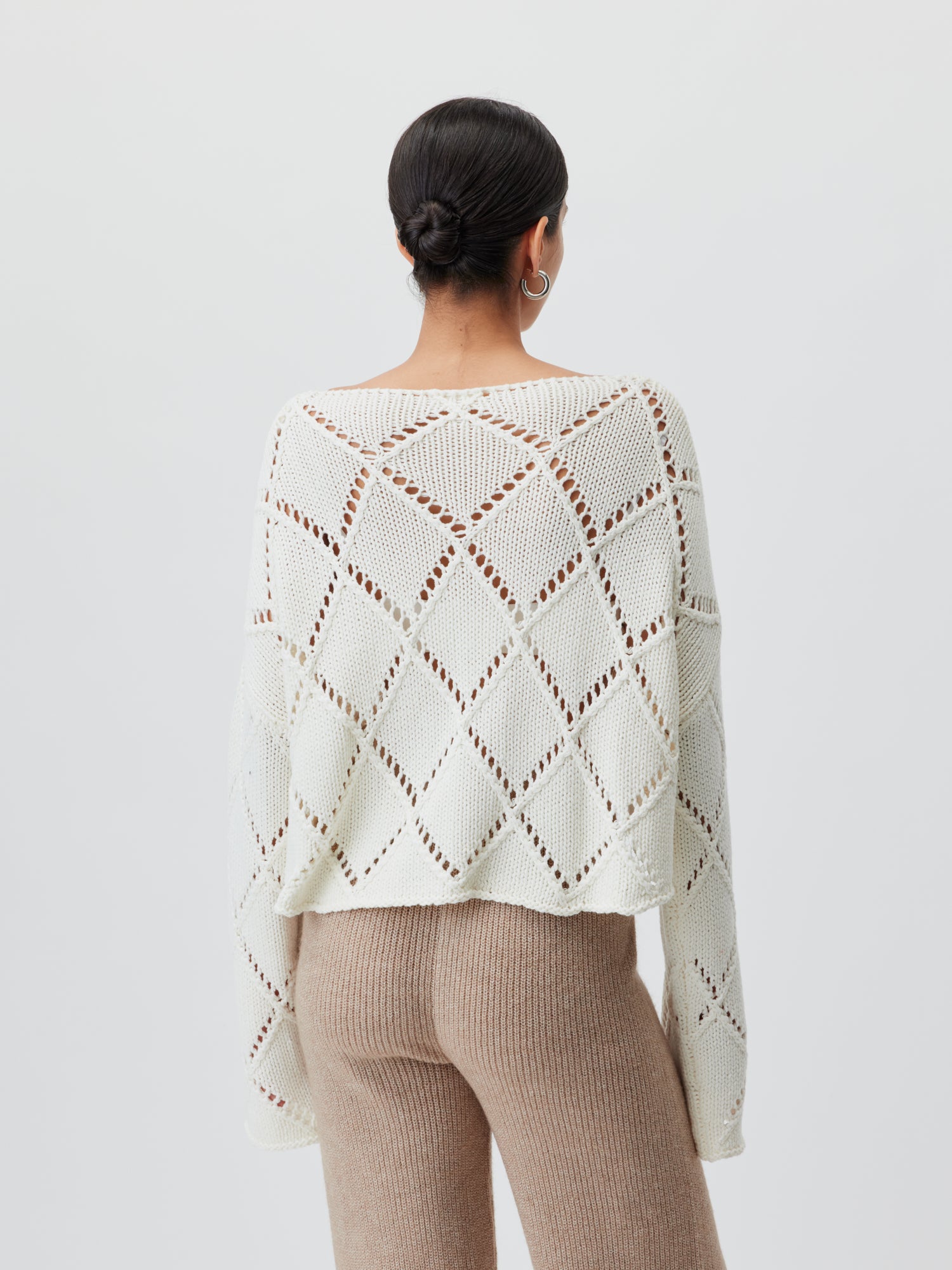 Eine Person ist von hinten zu sehen und trägt den Briley Jumper, einen lockeren, weißen Strickpullover mit Rautenmuster von LeGer by Lena Gercke, kombiniert mit langen beigen Strickhosen. Mit zu einem Dutt gestyltem dunklen Haar zeigt sie die Passform ihres Outfits vor einem schlichten hellen Hintergrund.