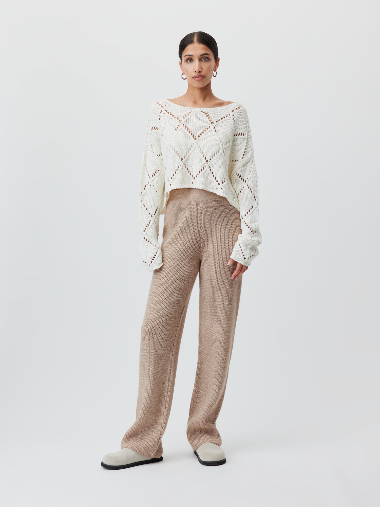 Eine Person steht vor einem weißen Hintergrund und trägt den Briley Jumper von LeGer by Lena Gercke, der ein offenes Strickdesign mit Rautenmuster und langen Ärmeln aufweist. Dazu trägt sie beige Strickhosen normaler Länge und cremefarbene Schuhe. Die Haare sind zurückgekämmt.