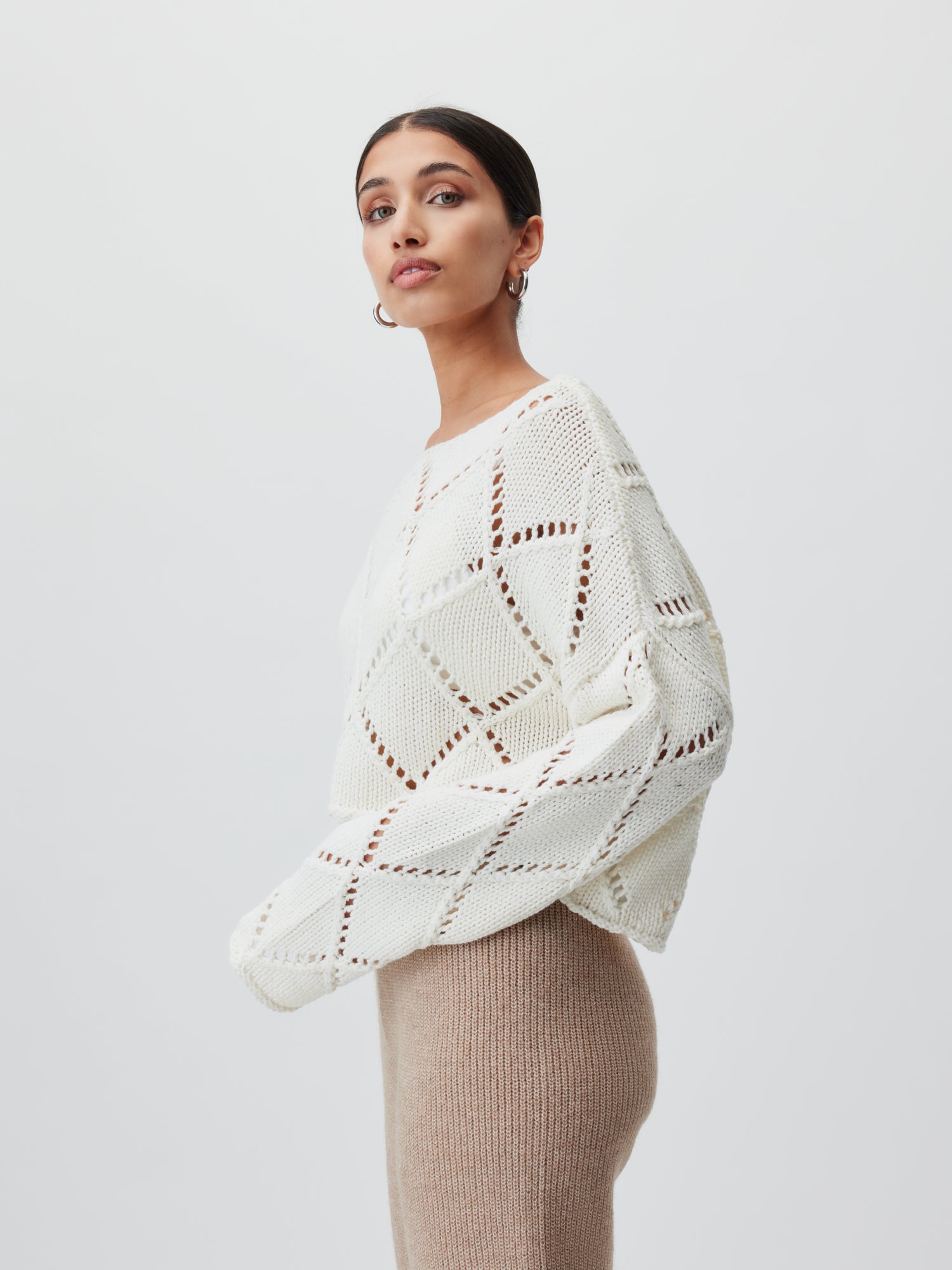 Eine Person präsentiert den Briley-Pullover von LeGer by Lena Gercke, ein weißes Strickmuster mit Rautenmuster, kombiniert mit einem beigen Rock. Ihr dunkles Haar ist zurückgekämmt und sie trägt Creolen vor einem schlichten weißen Hintergrund.