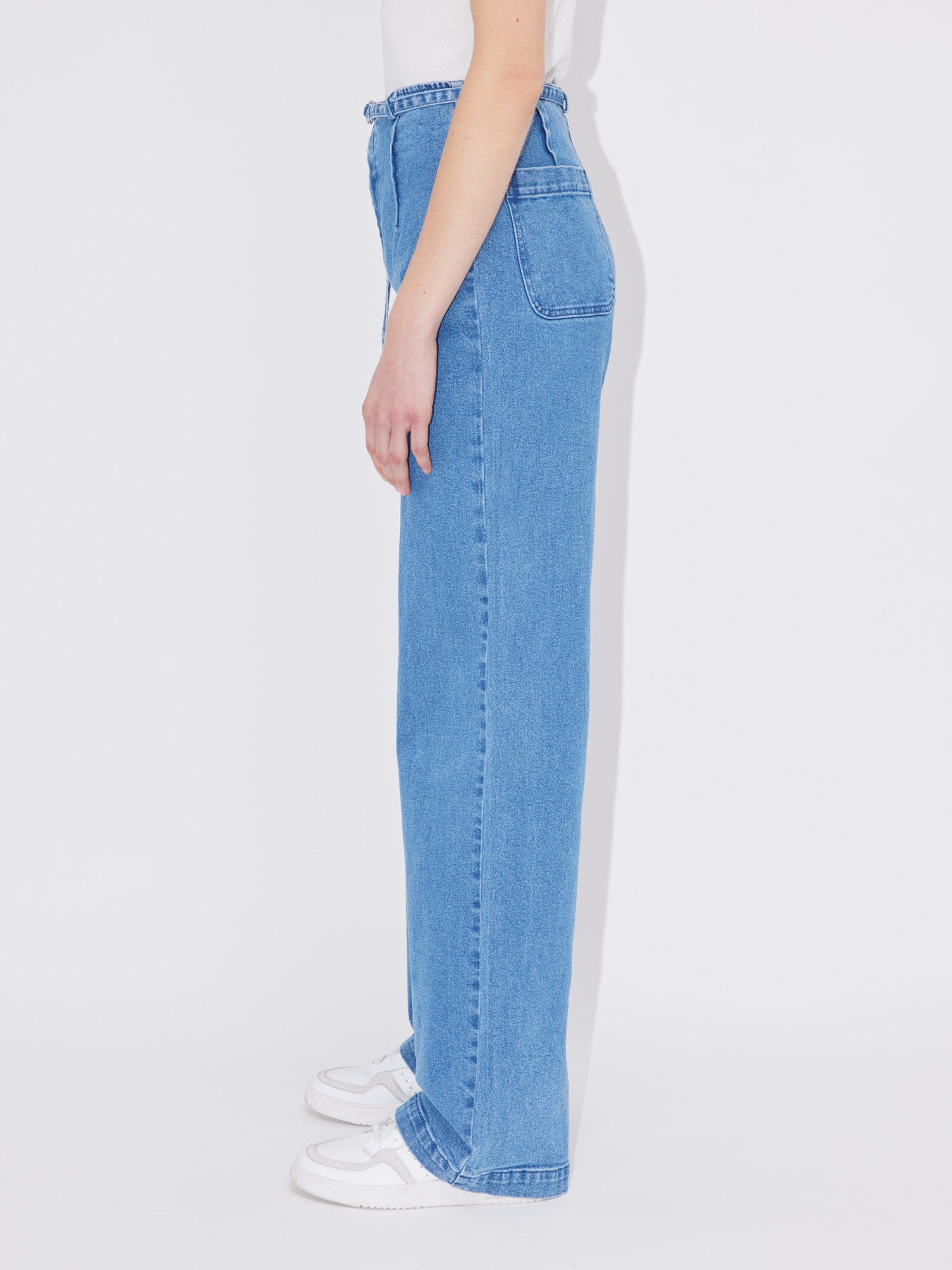 Eine Person steht im Profil und trägt die hoch taillierte, locker sitzende „Nanni Tall“-Bluejeans von LeGer by Lena Gercke und weiße Sneakers. Das Oberteil, das in der Länge Lang/Maxi zu sein scheint, ist hineingesteckt, aber nicht vollständig sichtbar. Der Hintergrund ist schlicht weiß.