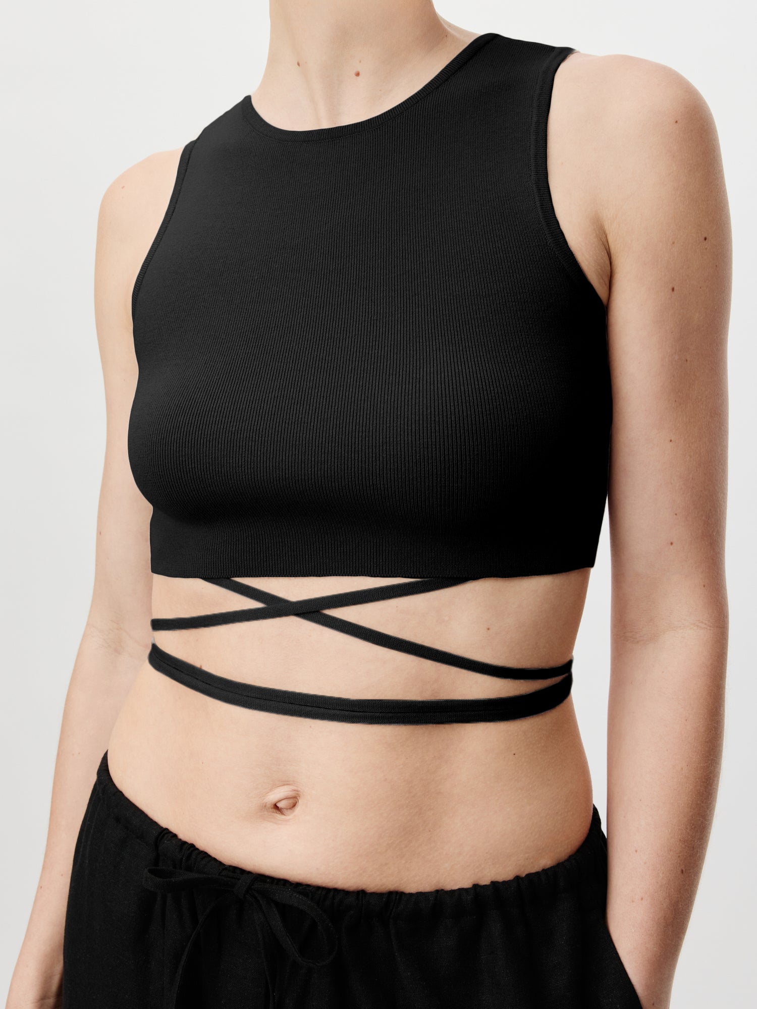 Person trägt das Top „Leany“ von LeGer by Lena Gercke, ein schwarzes ärmelloses Crop-Top mit überkreuzten Trägern, hohem Ausschnitt und gerippter Textur. Kombiniert mit einer schwarzen Hose unterstreicht das figurbetonte Design den eleganten Stil.