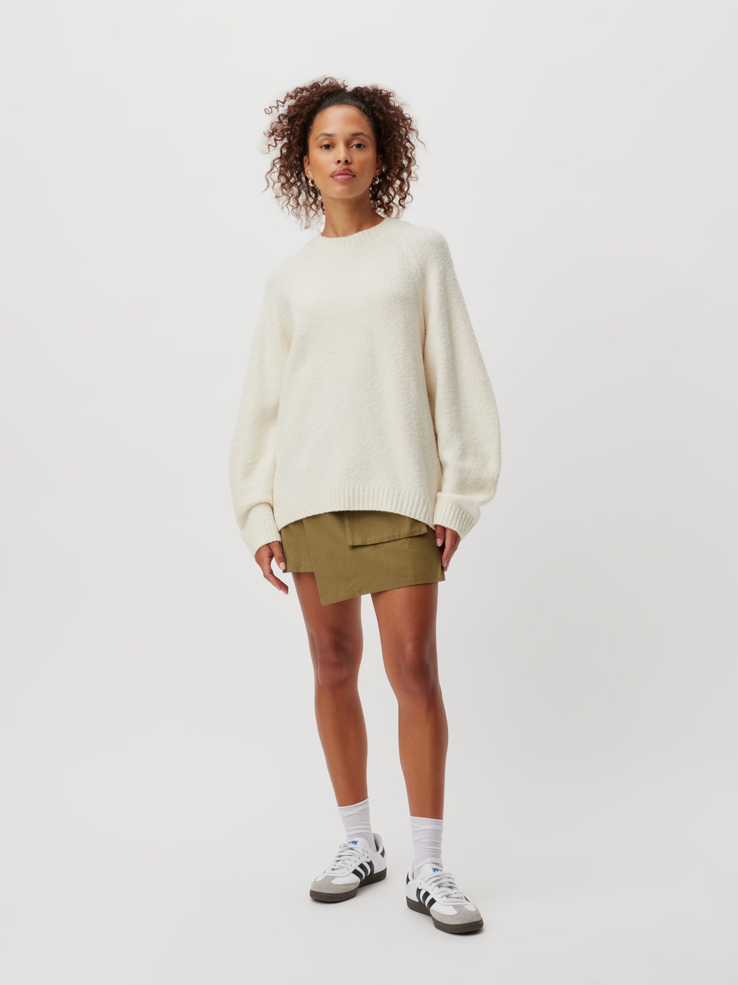 Eine Frau mit lockigem Haar trägt den Pullover Elmira von LeGer by Lena Gercke in Creme, kombiniert mit olivgrünen Shorts. Sie steht vor einem schlichten weißen Hintergrund, ergänzt durch weiße Socken und weiße Sneakers mit schwarzen Streifen. Die Passform des Outfits sorgt für einen bequemen und stilvollen Look.