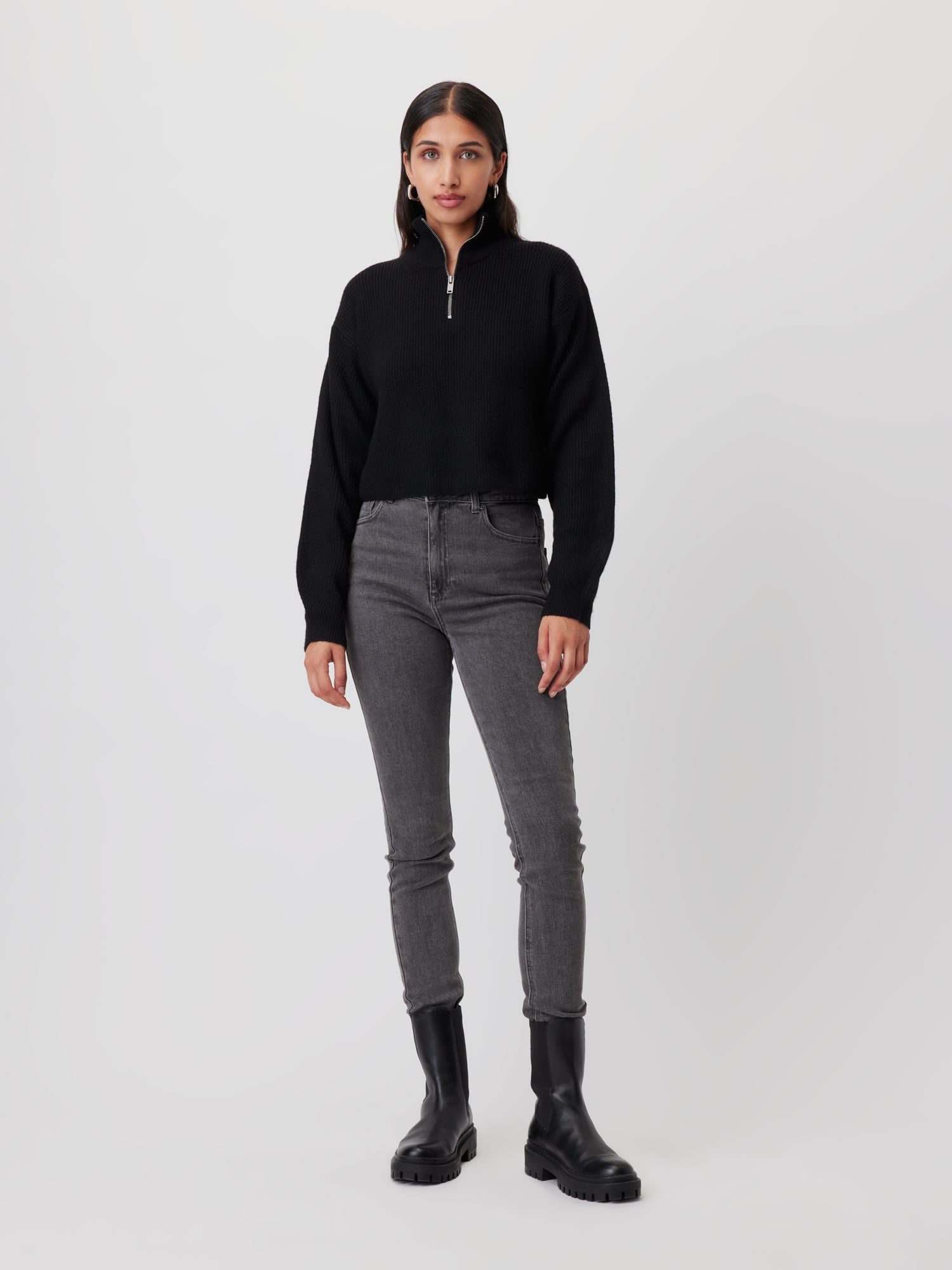 Eine Frau mit langen dunklen Haaren steht vor einem schlichten Hintergrund. Sie trägt einen schwarzen Pullover „Melissa“ mit Reißverschluss von LeGer by Lena Gercke, graue Skinny Jeans mit hoher Taille und schwarze Stiefeletten. Sie blickt in die Kamera, ihre Arme ruhen an den Seiten und zeigen die Passform ihres stylischen Outfits.