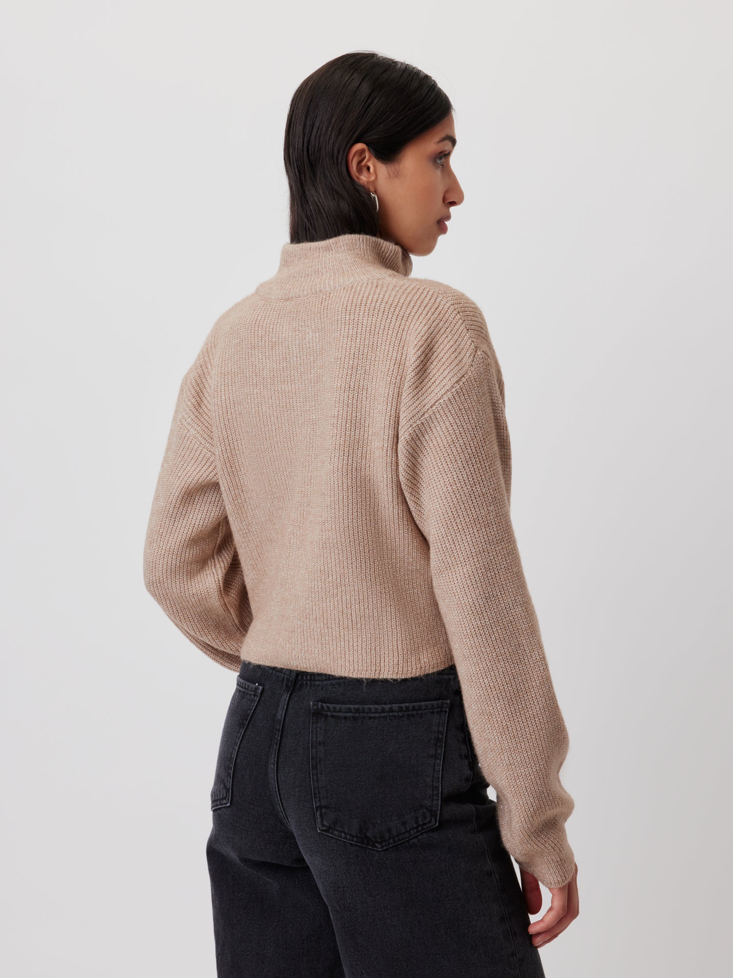 Eine Person mit langen dunklen Haaren trägt von hinten betrachtet den beigefarbenen Rollkragenpullover „Melissa“ mit Langarm-Design von LeGer by Lena Gercke und schwarze Jeans. Die Passform des Pullovers schmeichelt ihrer Figur perfekt, während sie vor einem schlichten weißen Hintergrund steht.