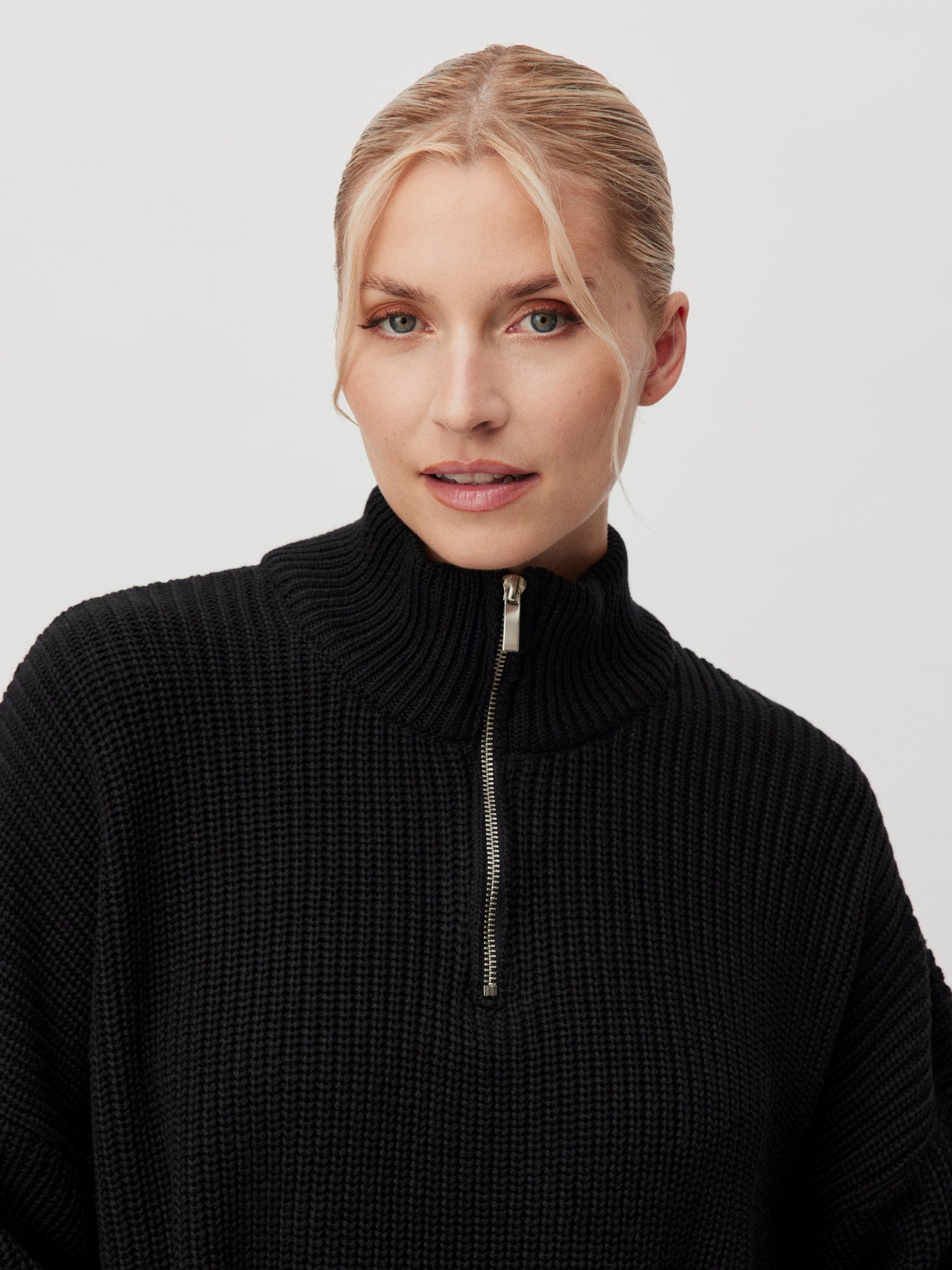 Eine Person mit blondem Haar trägt den Pullover „Emma“, einen hochgeschlossenen schwarzen Pullover mit einem halben Reißverschluss vorne. Das Langarm-Design des von LeGer by Lena Gercke entworfenen Pullovers ergänzt seine normale Länge und Passform. Sie blickt mit neutralem Gesichtsausdruck vor einem schlichten weißen Hintergrund direkt in die Kamera.