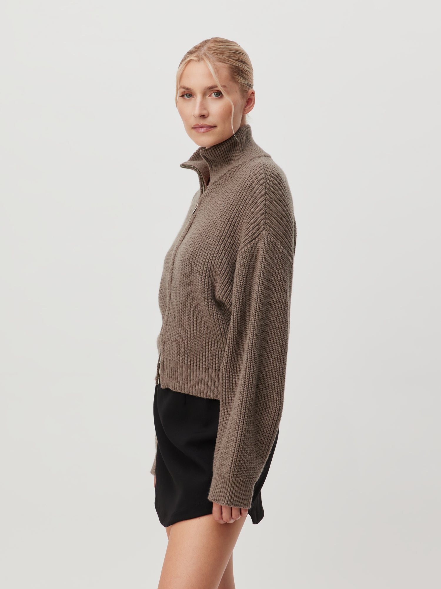 Eine Person mit blonden Haaren posiert vor einem schlichten Hintergrund. Sie trägt einen taupefarbenen, langärmeligen Rippenpullover mit hohem Kragen und normaler Länge sowie die Strickjacke „Cindy“ von LeGer by Lena Gercke, kombiniert mit einem schwarzen Rock. Die Person ist leicht zur Seite gewandt und blickt mit ruhigem Gesichtsausdruck in die Kamera.