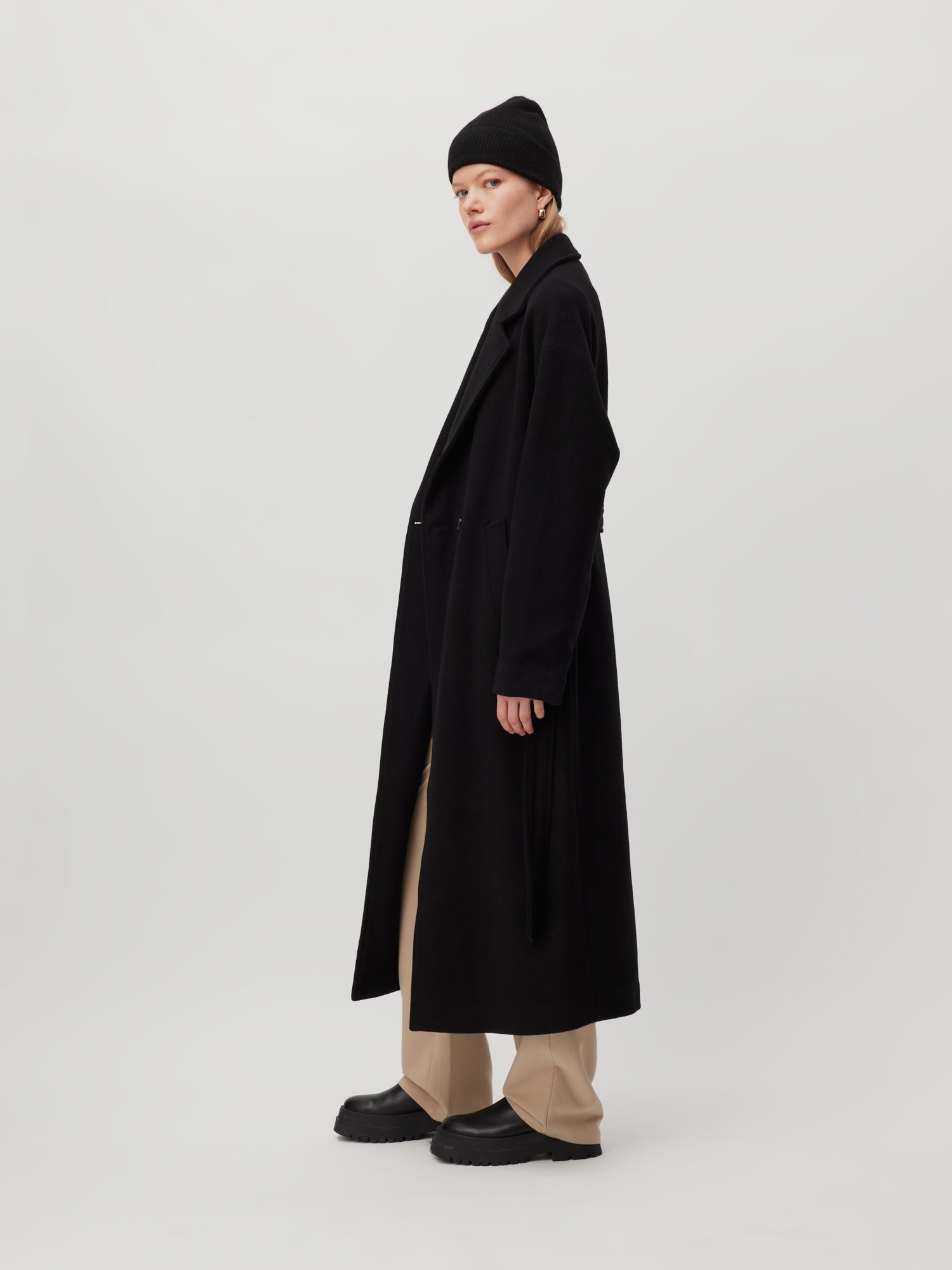 Eine Person, die den Dena Coat von LeGer by Lena Gercke trägt, der sich durch seine optimale Passform und sein langes schwarzes Design auszeichnet, steht vor einem hellgrauen Hintergrund. Sie kombiniert ihn mit beigen Hosen und einer schwarzen Beanie und blickt mit neutralem Gesichtsausdruck nach links.