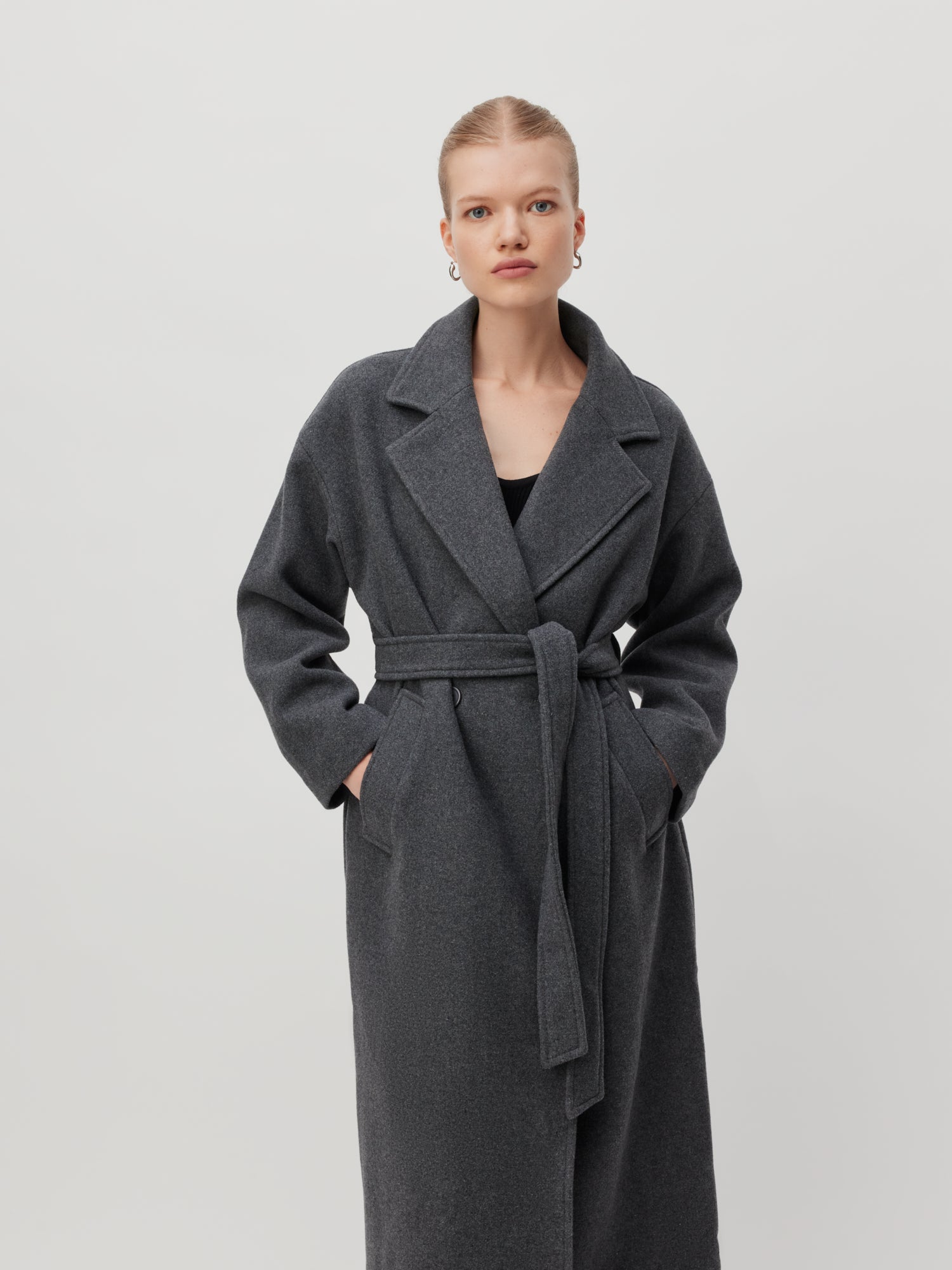 Eine Person mit kurzen blonden Haaren trägt den langen, grauen Dena Coat von LeGer by Lena Gercke. Die Hände in den Manteltaschen vergraben, steht sie vor einem schlichten, hellen Hintergrund. Ihr stilvolles Ensemble mit Creolen bietet eine normale Passform und normale Länge für einen klassischen Look.