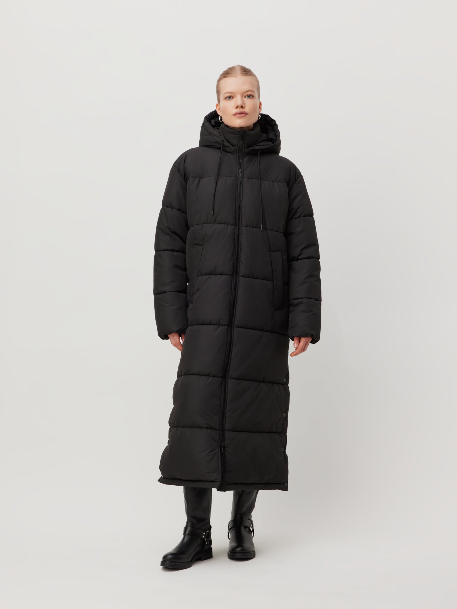 Eine Person trägt den Donia Coat von LeGer by Lena Gercke, einen langen schwarzen Puffermantel mit Kapuze und großen Taschen, vor einem schlichten Hintergrund. Das Langarm-Design des Mantels unterstreicht seinen stilvollen Reiz. Passend dazu vervollständigen sie den Look mit schwarzen Stiefeln.