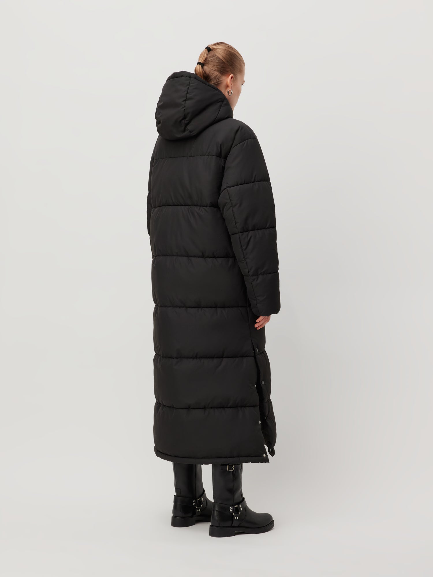 Eine Person trägt den Donia Coat von LeGer by Lena Gercke, einen langen schwarzen Puffermantel mit Kapuze, der sein knielanges Design betont, während sie mit dem Gesicht nach außen steht. Schwarze Stiefel runden dieses elegante Ensemble perfekt ab. Der schlichte, helle Hintergrund verstärkt den schlichten, aber modischen Reiz dieses Outfits.