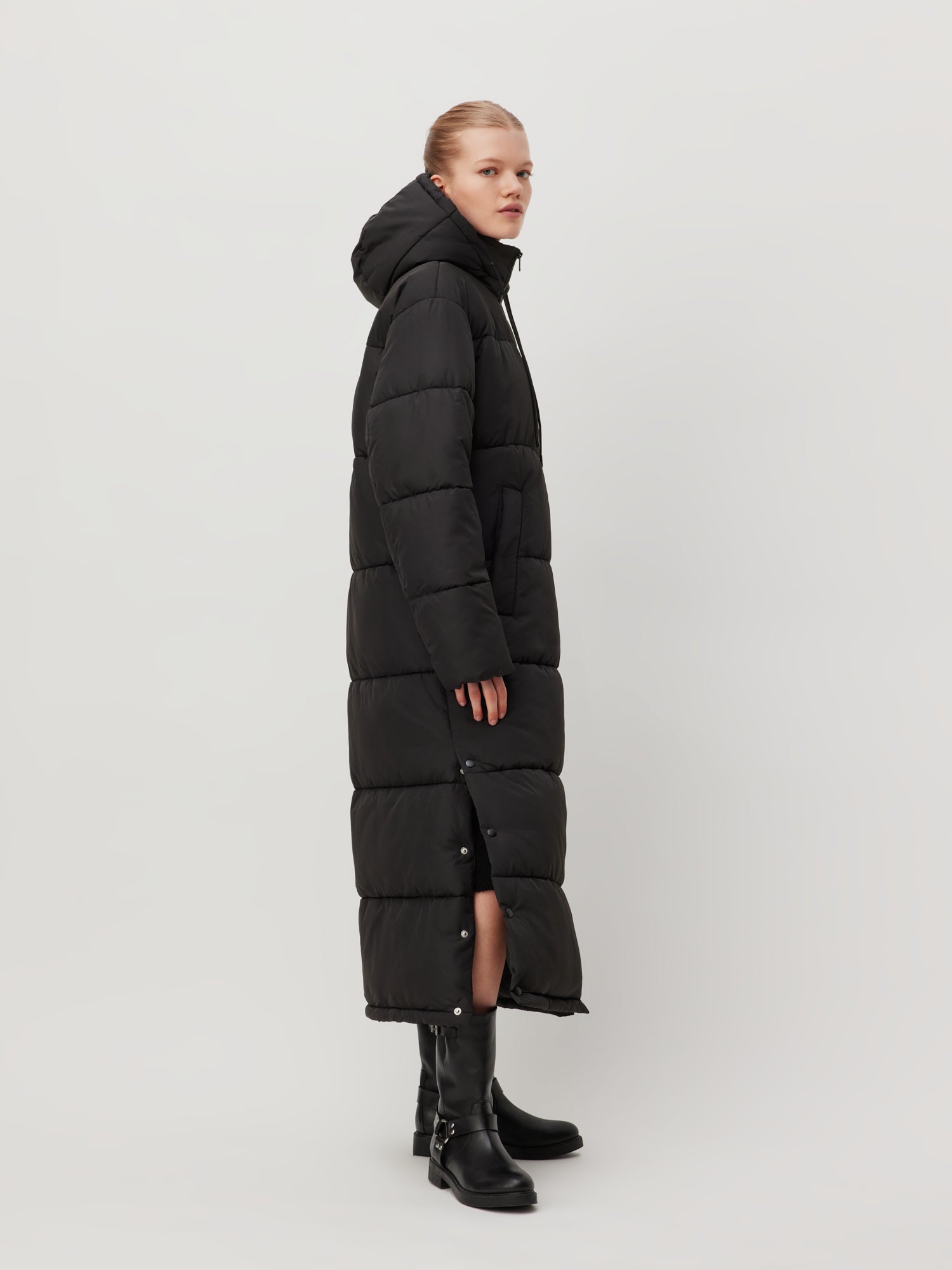 Eine Person ist im Seitenprofil zu sehen und trägt den Donia Coat von LeGer by Lena Gercke, ein langes, schwarzes Puffer-Design mit langen Ärmeln und einer Kapuze, kombiniert mit schwarzen Stiefeln. Der Mantel ist auf seiner Standardlänge zugeknöpft und steht vor einem schlichten, hellen Hintergrund.