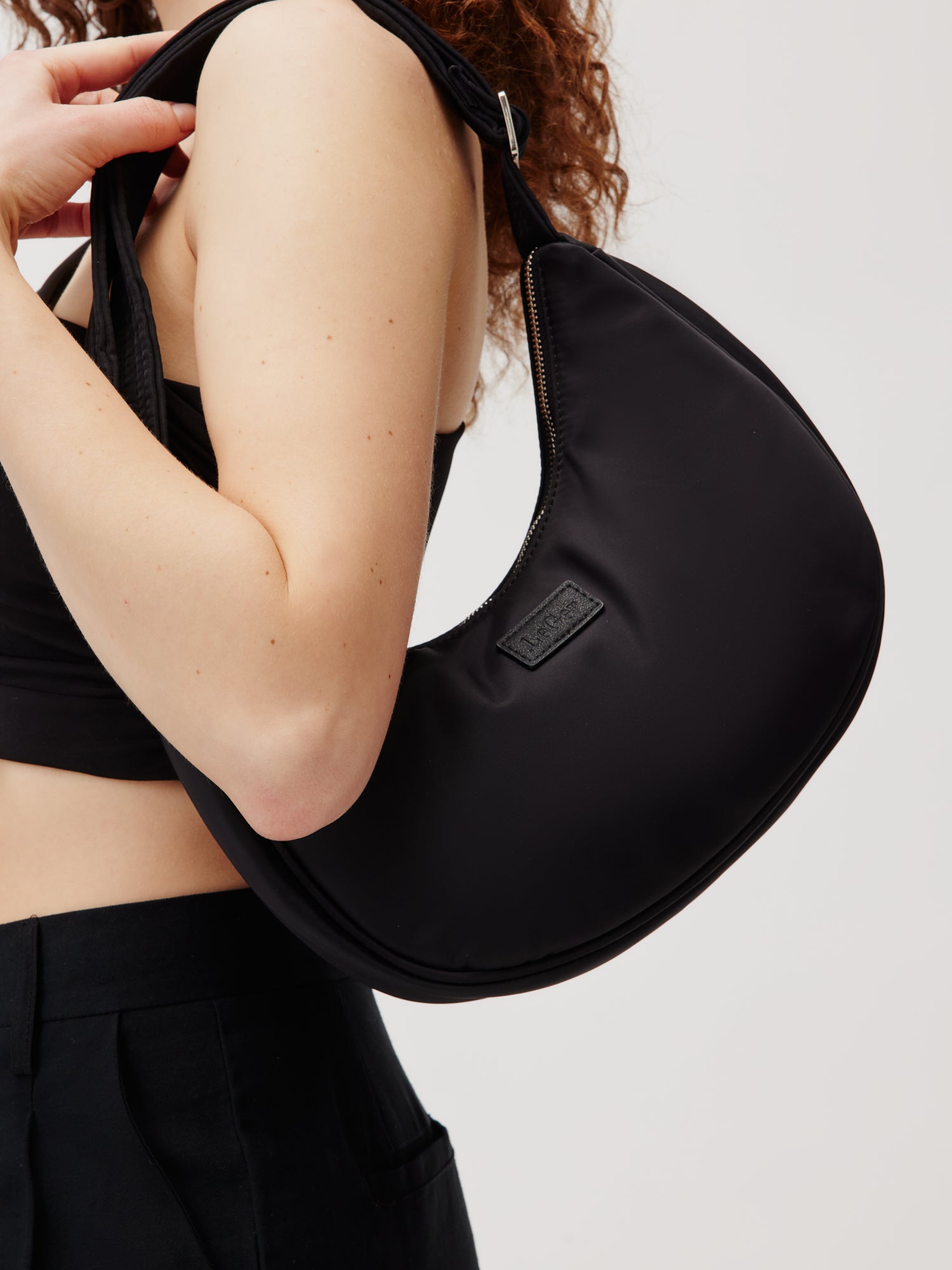 Eine Person mit lockigem Haar hält die schicke Bora Bag von LeGer by Lena Gercke und präsentiert ihr schlichtes Design und den Logo-Aufnäher. Die stilvolle schwarze, halbmondförmige Umhängetasche verfügt über einen Reißverschluss und akzentuiert ihr schwarzes Oberteil und ihre Hose vor einem neutralen Hintergrund. Dieses Produkt unterstreicht perfekt die schlichte Modeästhetik und Funktionalität.