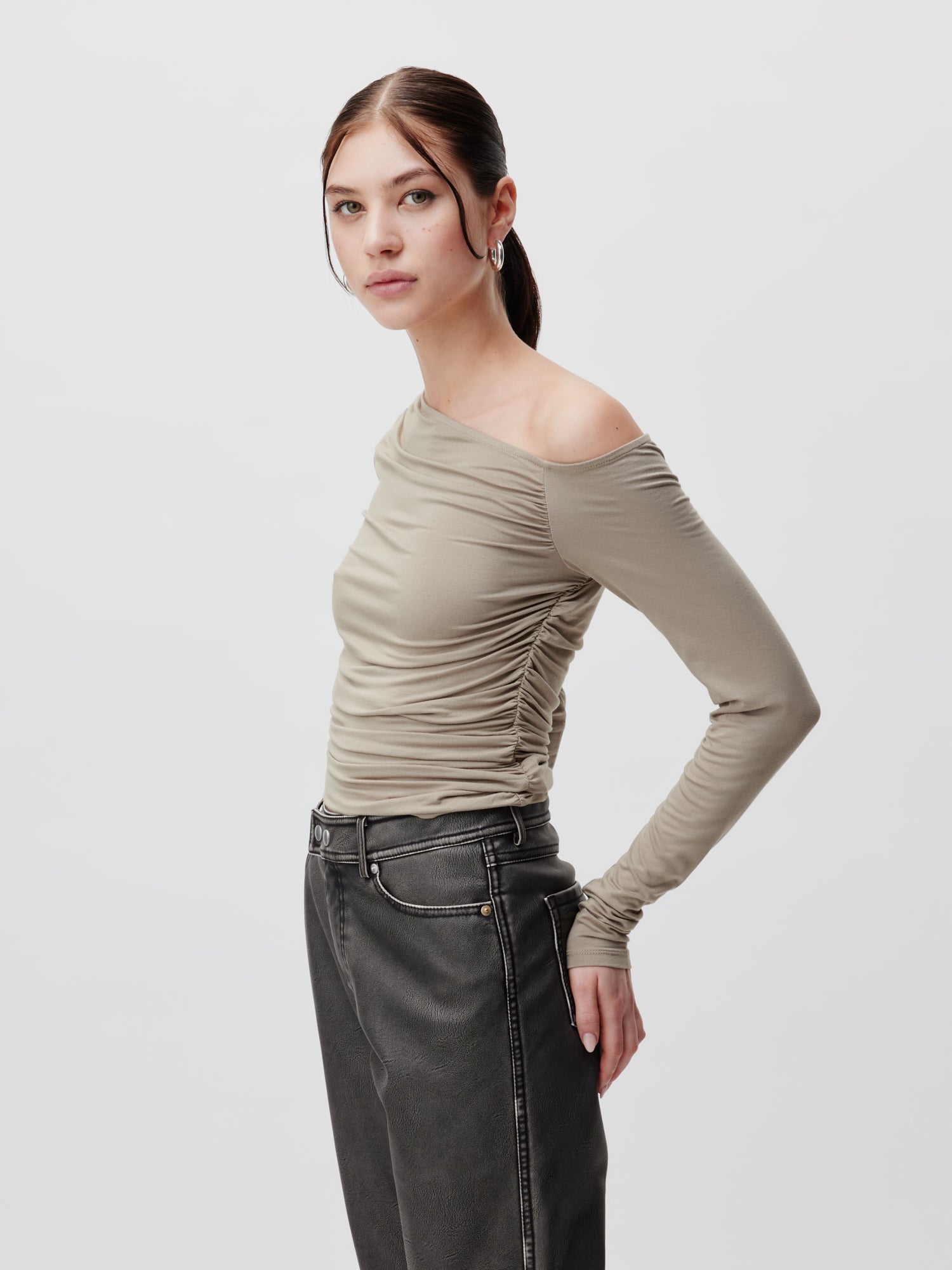 Eine Frau mit langen braunen Haaren trägt das Shirt „Inka“ von LeGer by Lena Gercke, ein graues One-Shoulder-Top mit gerüschten Details und dunkle Jeans mit hoher Taille. Das Oberteil hat eine Passform, die ihre Figur betont. Sie blickt in die Kamera und steht vor einem neutralen hellgrauen Hintergrund.