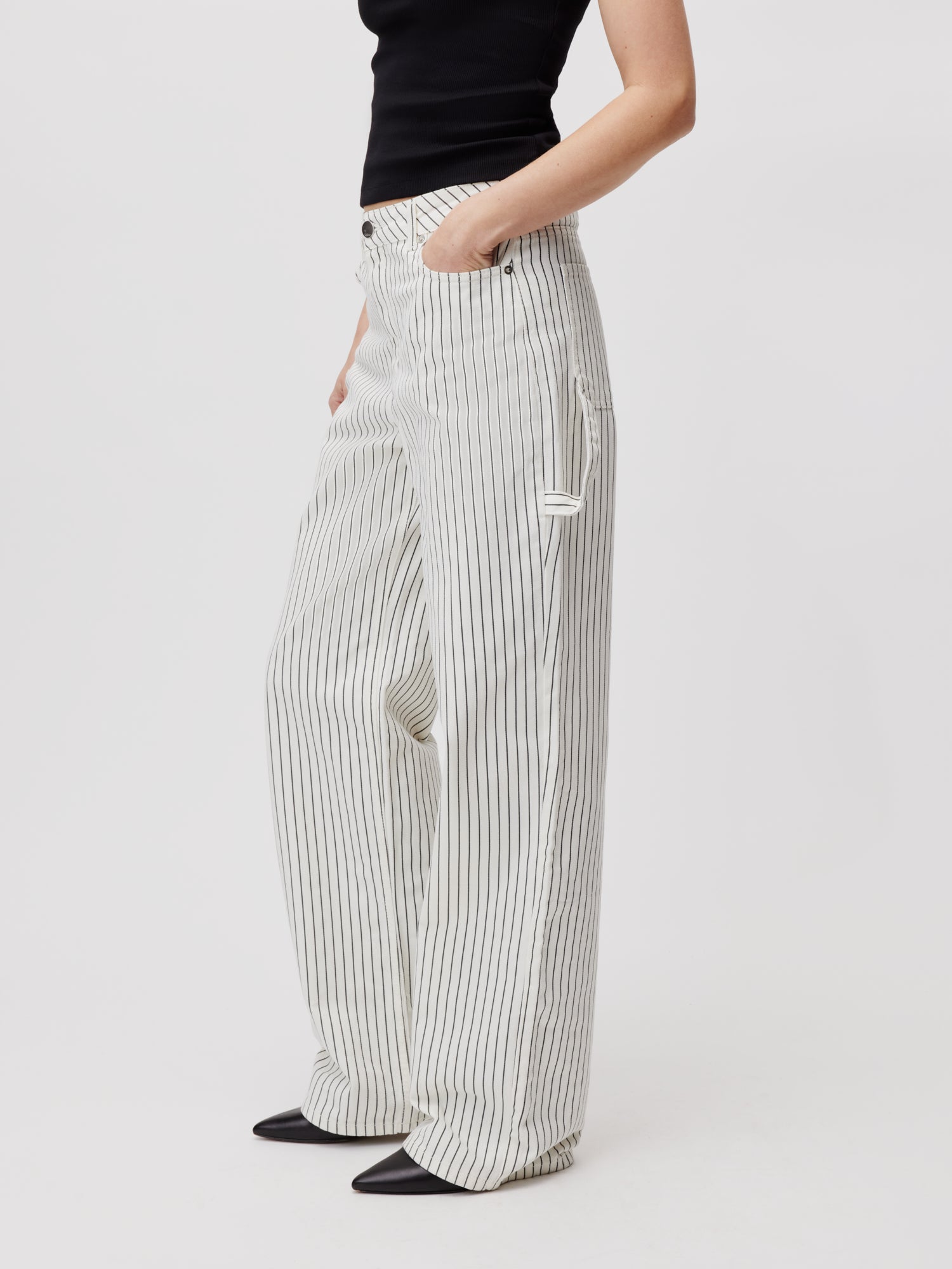 Eine Person, die ein schwarzes ärmelloses Oberteil und die Lisanna Pants Tall von LeGer by Lena Gercke trägt – ein weißes, hoch tailliertes, weites Design mit dünnen vertikalen Streifen – nimmt eine selbstbewusste Pose ein. Ihre rechte Hand ruht lässig in der Tasche dieser stylischen Maxihose und hebt sich deutlich vom schlichten weißen Hintergrund ab.