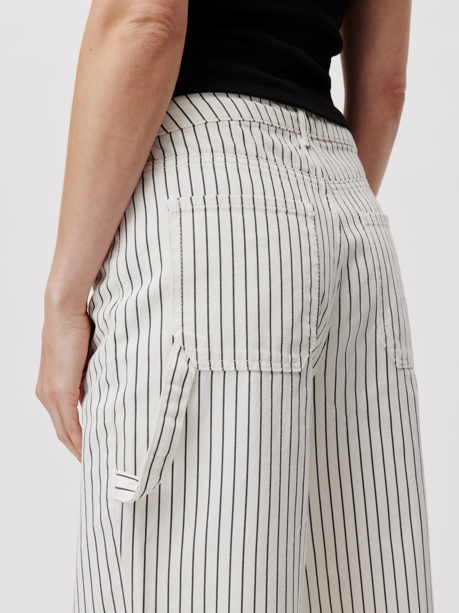 Ein Model trägt von der Taille abwärts die Lisanna Pants Tall von LeGer by Lena Gercke. Die locker geschnittene, schwarz-weiß gestreifte Hose hat zwei Gesäßtaschen und wird mit einem schwarzen Oberteil kombiniert, alles vor einem schlichten weißen Hintergrund.