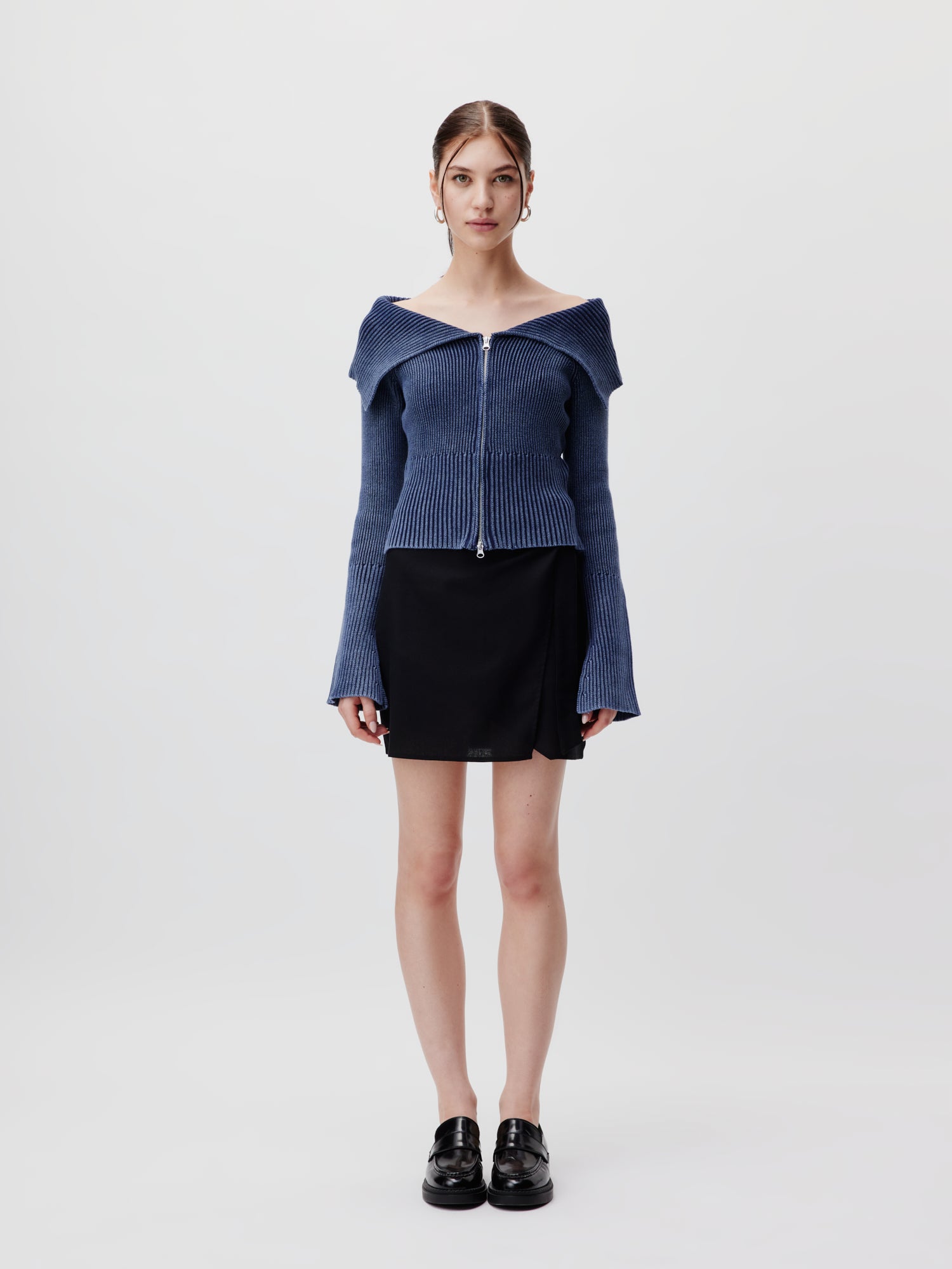 Eine Person steht vor einem schlichten Hintergrund und trägt den Anaida Cardigan von LeGer by Lena Gercke. Dieser Cardigan ist schulterfrei mit Reißverschluss und mit einem schwarzen Rock kombiniert. Er hat eine lange Passform und normale Länge und passt zu ihren schulterlangen Haaren und schwarzen Schuhen.