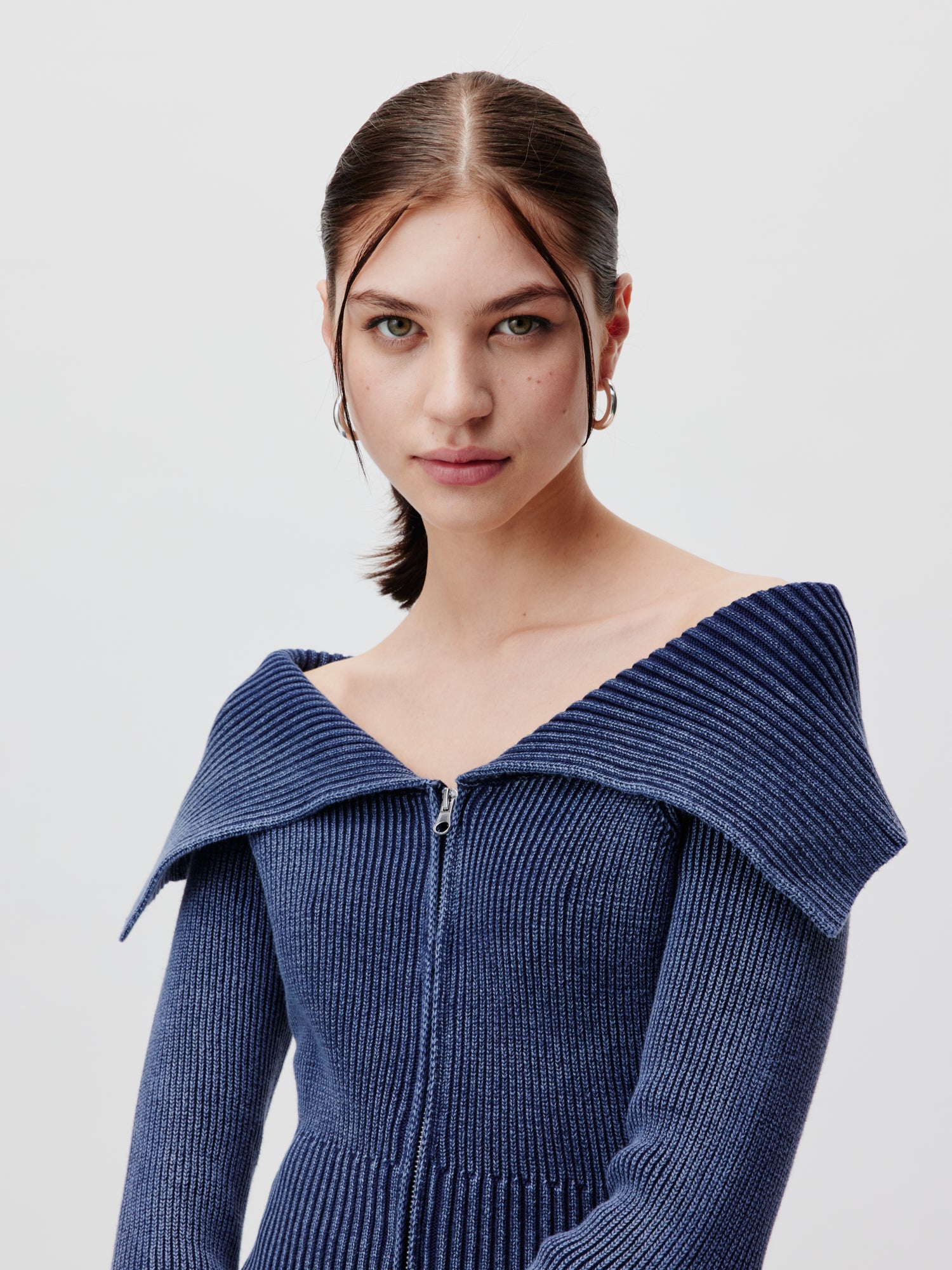 Eine Person mit zurückgebundenen langen dunklen Haaren trägt den Anaida Cardigan von LeGer by Lena Gercke, ein blaues, geripptes Off-Shoulder-Top mit Frontreißverschluss und Langarm-Design. Sie blickt vor einem schlichten hellen Hintergrund direkt in die Kamera.