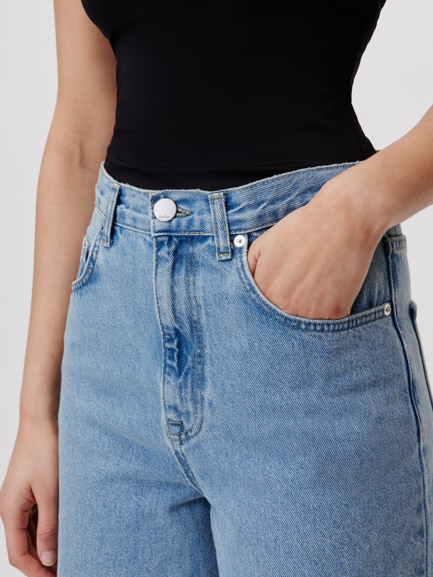 Eine Person trägt ein schwarzes Oberteil und die stylische Jeans „Cleo Tall“ von LeGer by Lena Gercke, die mit einer hohen Taille für eine perfekte Passform entworfen wurde. Die Jeans hat silberne Knopfdetails und auffällige Nähte. Ihre rechte Hand steckt lässig in der Tasche, vor einer schlichten weißen Wand.