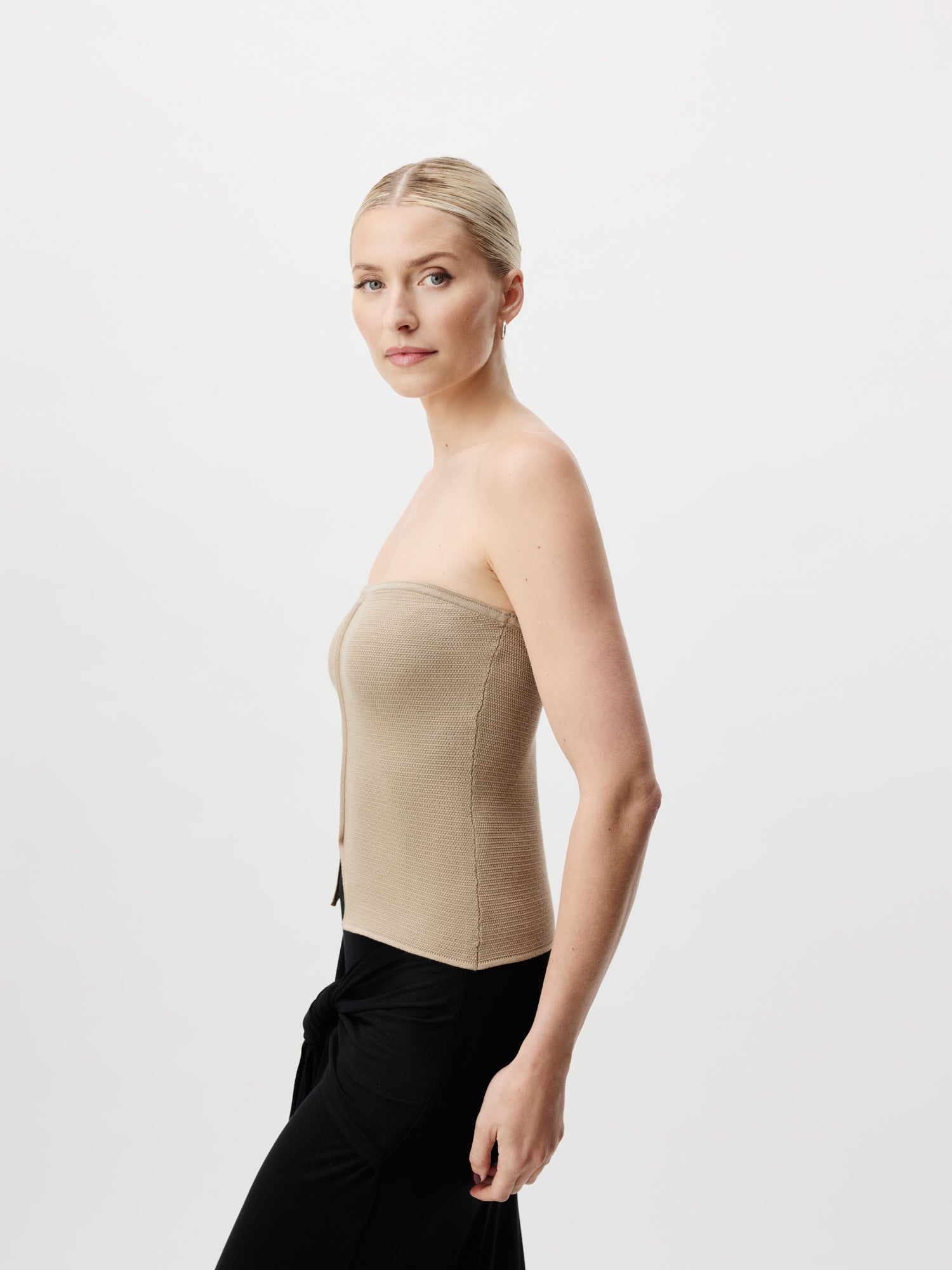 Eine Frau mit zurückgekämmten blonden Haaren posiert elegant vor einem schlichten weißen Hintergrund. Sie trägt das schulterfreie Fabiola Top in Beige von LeGer by Lena Gercke und eine schmale schwarze Hose. Im Profil stehend blickt sie selbstbewusst in die Kamera.