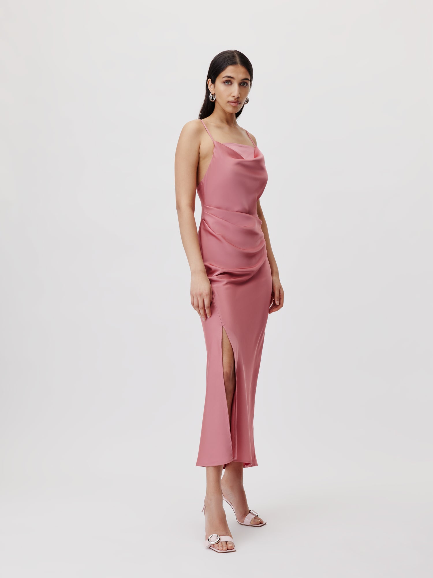 Eine Frau steht vor einem schlichten Hintergrund und trägt das Brianne-Kleid von LeGer by Lena Gercke, ein rosa Maxikleid aus Satin mit oberschenkelhohem Schlitz und ärmellosem Design. Sie hat langes dunkles Haar und ergänzt ihren Look mit Ohrringen und offenen High Heels.