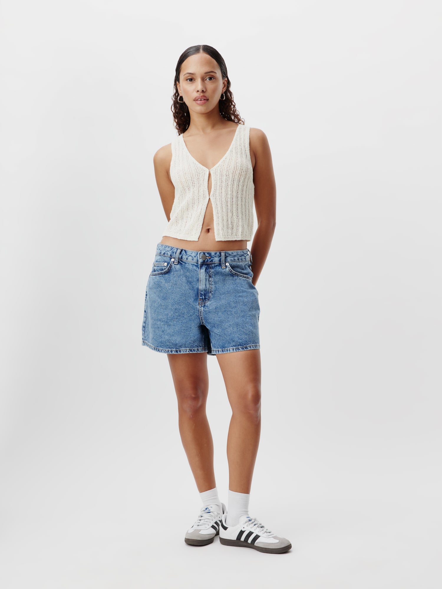 Eine Person steht vor einem schlichten weißen Hintergrund und trägt ein weißes ärmelloses Strickoberteil und die Nena-Shorts von LeGer by Lena Gercke in blauem Denim, ideal für alle, die Größe 36 suchen. Sie hat lange Haare, trägt weiße Socken und schwarz-weiße Turnschuhe und lässt die Hände locker an den Seiten hängen.