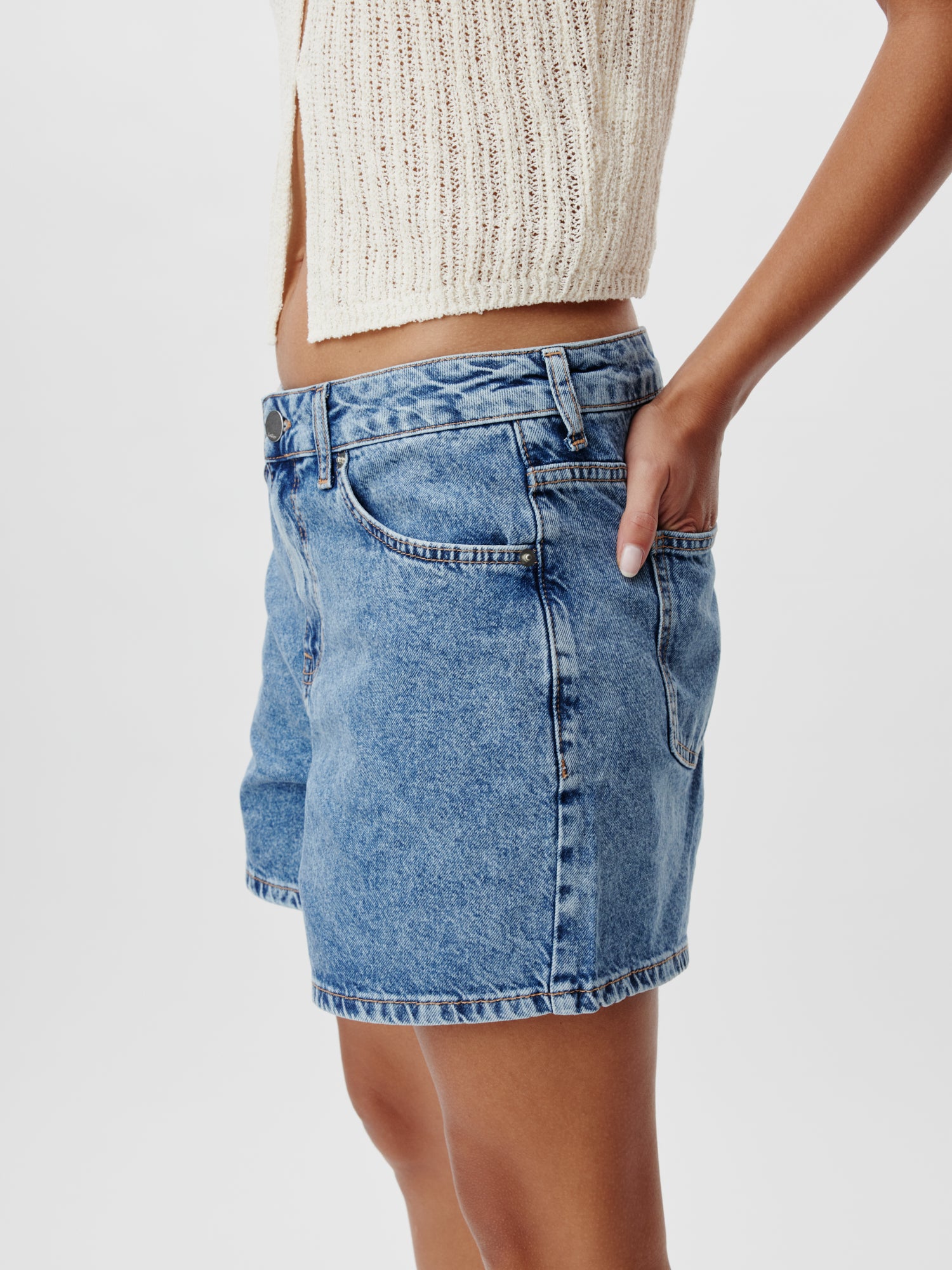 Eine Person trägt die hoch taillierten Nena-Shorts von LeGer by Lena Gercke in hellblauem Denim, Größe 36, und kombiniert sie mit einem cremefarbenen, kurzen Stricktop. Das stylische Kurz-/Mini-Ensemble wird durch eine Hand lässig in der Tasche vor einem schlichten weißen Hintergrund vervollständigt.