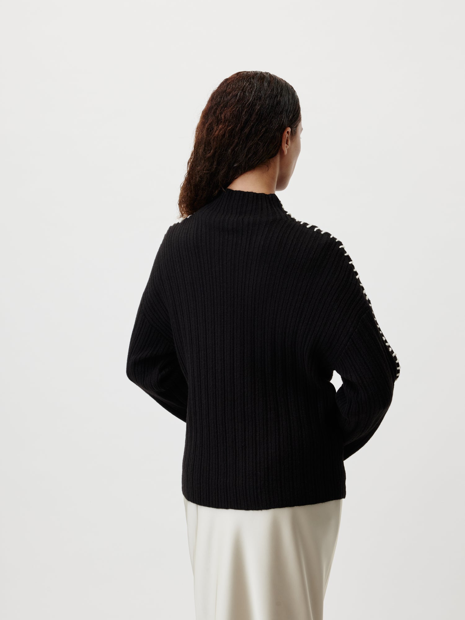 Eine Person mit langen lockigen Haaren ist von hinten zu sehen, sie trägt den Ronja Pullover von LeGer by Lena Gercke. Der Pullover ist schwarz, gerippt, hat normale Länge und lange Ärmel und sie steht vor einem schlichten weißen Hintergrund. Der untere Teil eines hellen Rocks oder einer hellen Hose ist zu sehen.