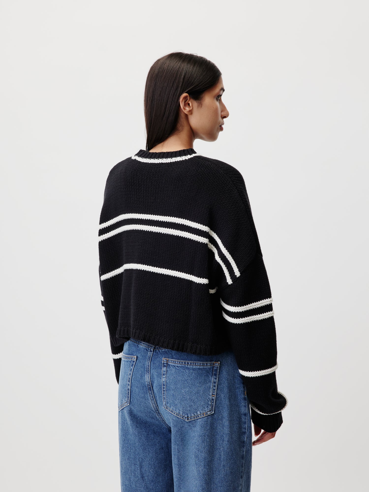 Eine Person mit langen dunklen Haaren steht seitlich und präsentiert den Ida Jumper von LeGer by Lena Gercke. Dieser schwarze Pullover hat weiße Querstreifen in einem langärmeligen und kurzen Design. Sie kombinieren ihn mühelos mit Blue Jeans auf einem schlichten, hellen Hintergrund.