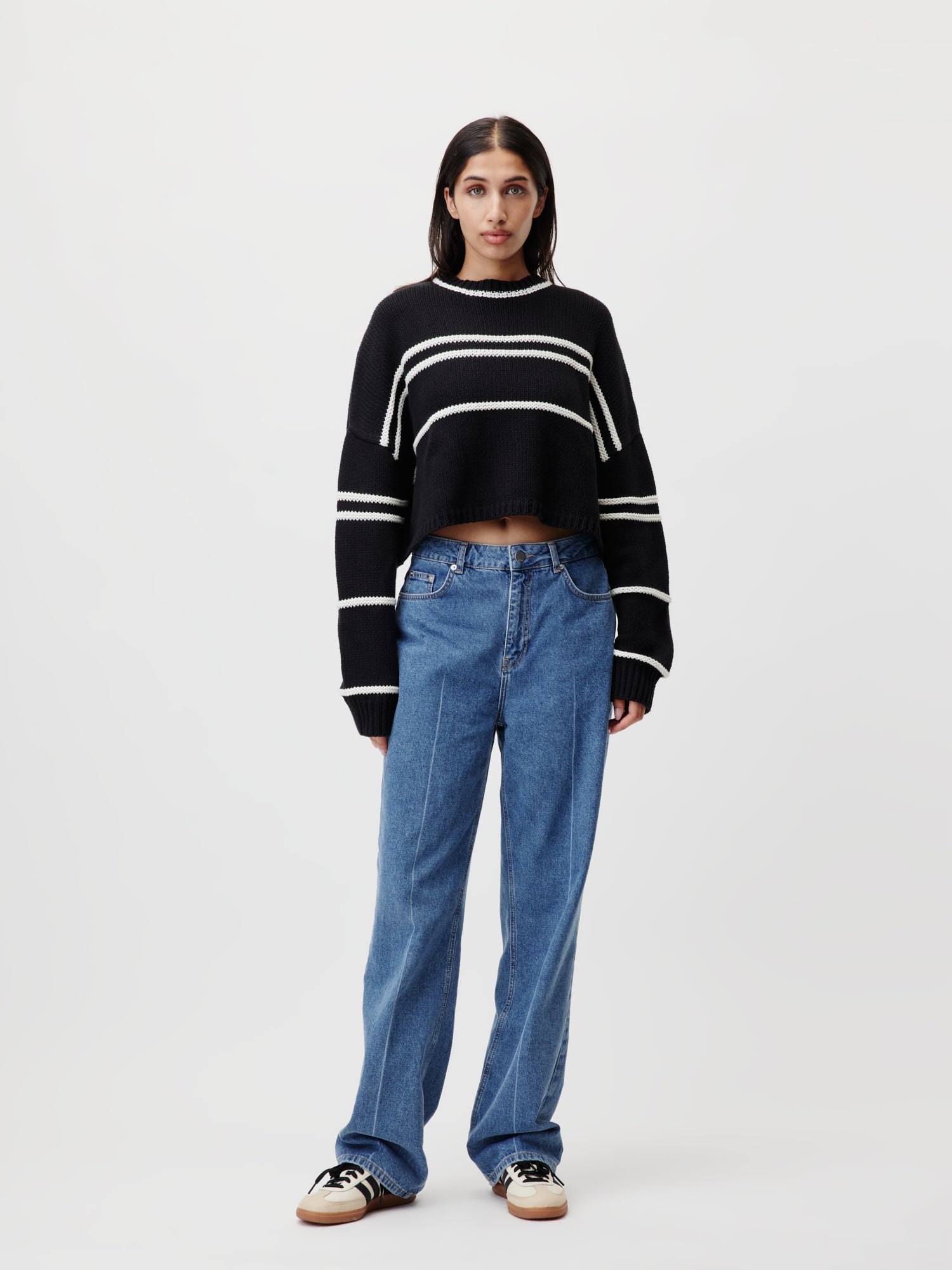 Eine Person steht vor einem schlichten Hintergrund und trägt den Ida-Pullover von LeGer by Lena Gercke mit schwarzen und weißen Streifen und kurzem Langarm-Design. Sie kombiniert ihn mit Bluejeans und schwarzen und weißen Sneakers, hat lange dunkle Haare und einen neutralen Gesichtsausdruck.