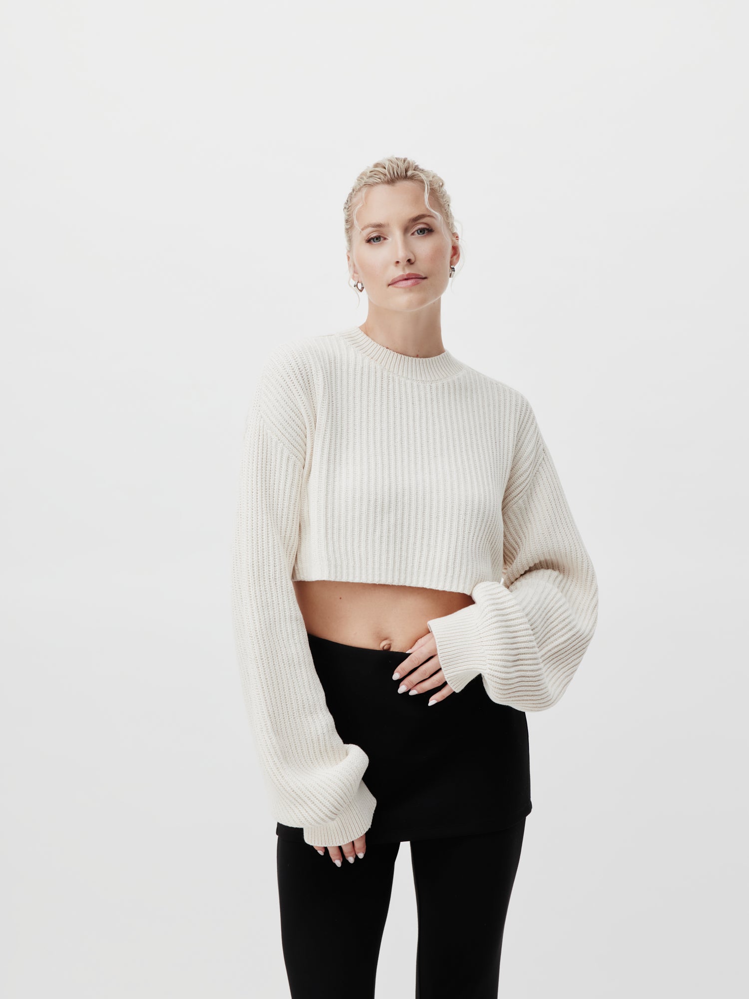 Eine Person mit kurzen blonden Haaren trägt den Thassia Jumper, einen cremefarbenen, gerippten Kurzpullover von LeGer by Lena Gercke, und einen schwarzen Rock. Sie steht vor einem schlichten weißen Hintergrund, hat eine Hand auf dem Bauch und einen ruhigen Gesichtsausdruck.