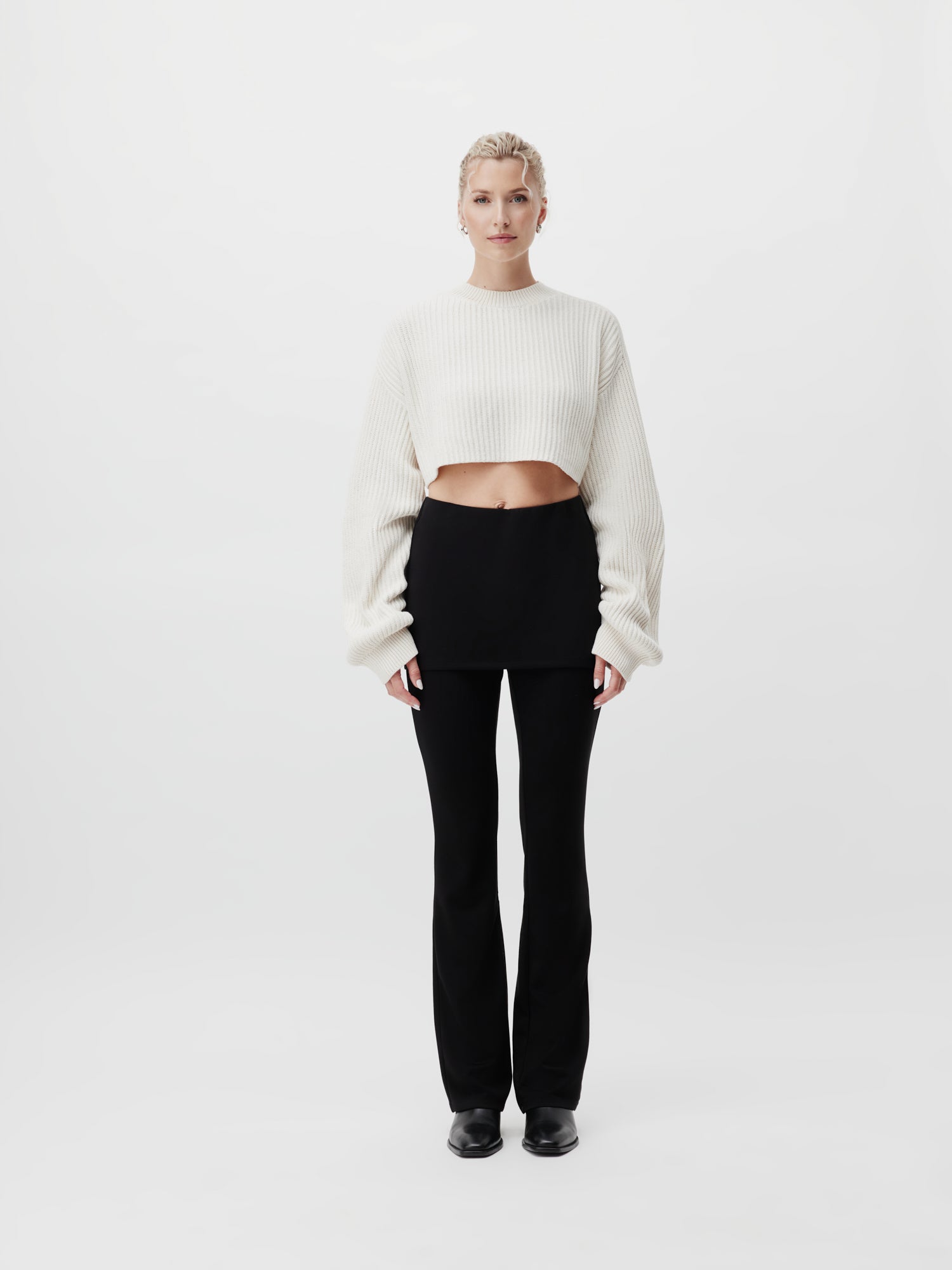 Eine Person steht vor einem schlichten weißen Hintergrund und präsentiert einen Thassia-Pullover von LeGer by Lena Gercke, kombiniert mit einem schwarzen Rock über einer passenden Hose und eleganten schwarzen Schuhen. Ihr Haar ist ordentlich gestylt. Das Outfit verkörpert eine minimalistische und monochrome Ästhetik mit einem kurzen Schnitt-Ansatz.