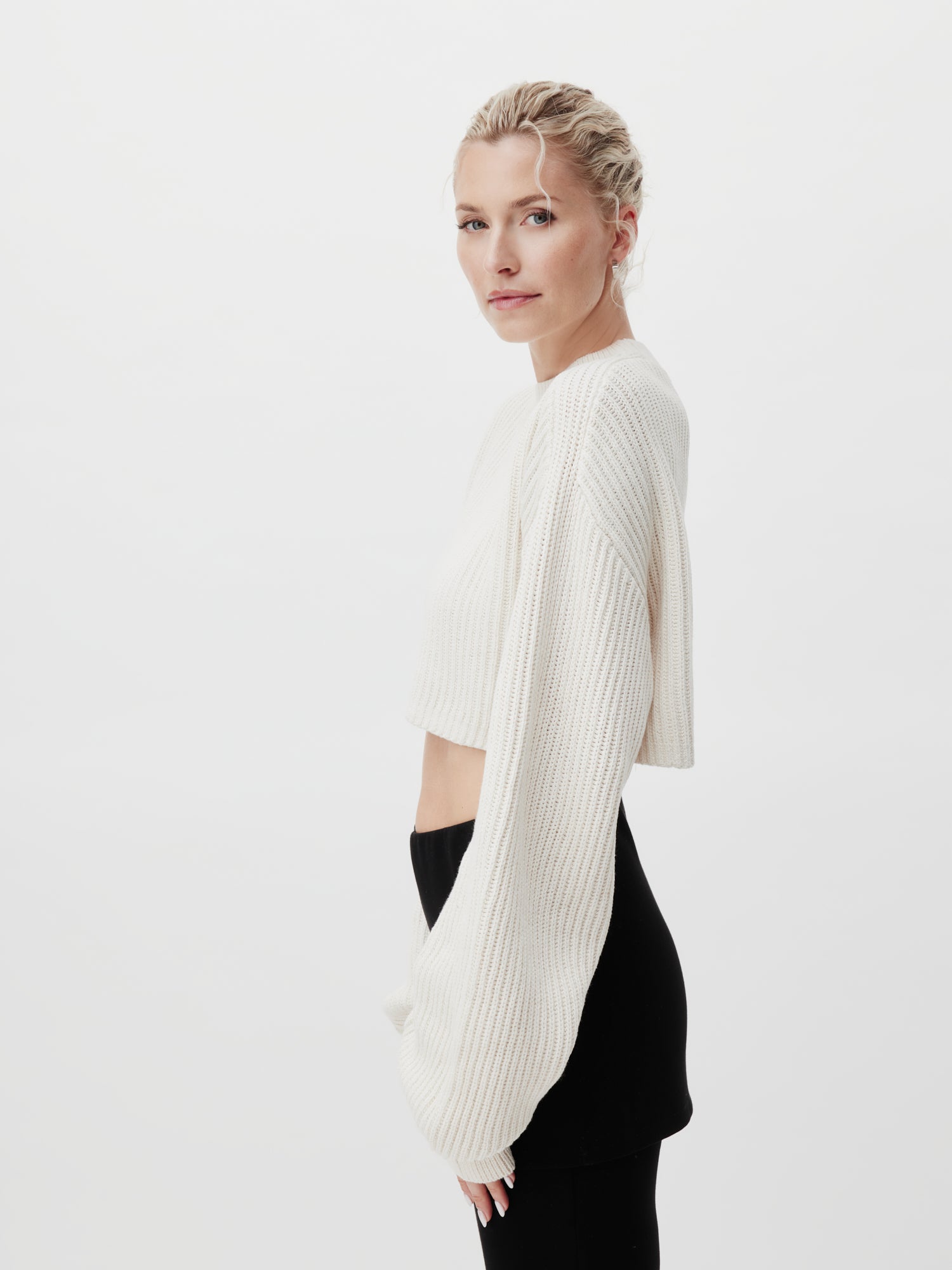 Eine Person mit blonden Haaren posiert vor einem weißen Hintergrund. Sie trägt den cremefarbenen Thassia-Pullover von LeGer by Lena Gercke im bauchfreien Top-Stil und eine schwarze Hose. Ihr Gesichtsausdruck ist neutral, da sie leicht zur Seite blickt.