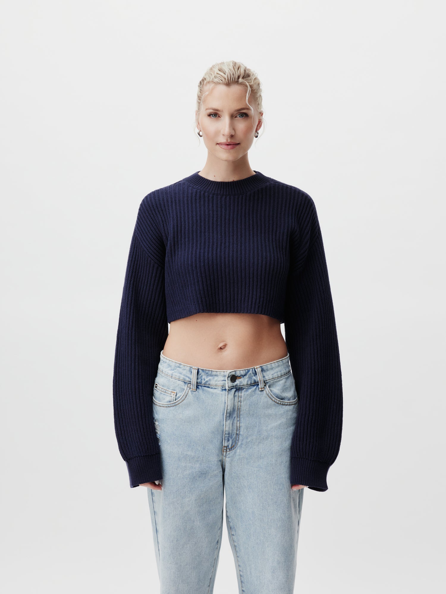 Eine Person mit kurzen blonden Haaren trägt den Pullover Thassia von LeGer by Lena Gercke im marineblauen Cropped-Style und hellblaue Jeans vor einem schlichten weißen Hintergrund und hebt das trendige Langarm-Design hervor.