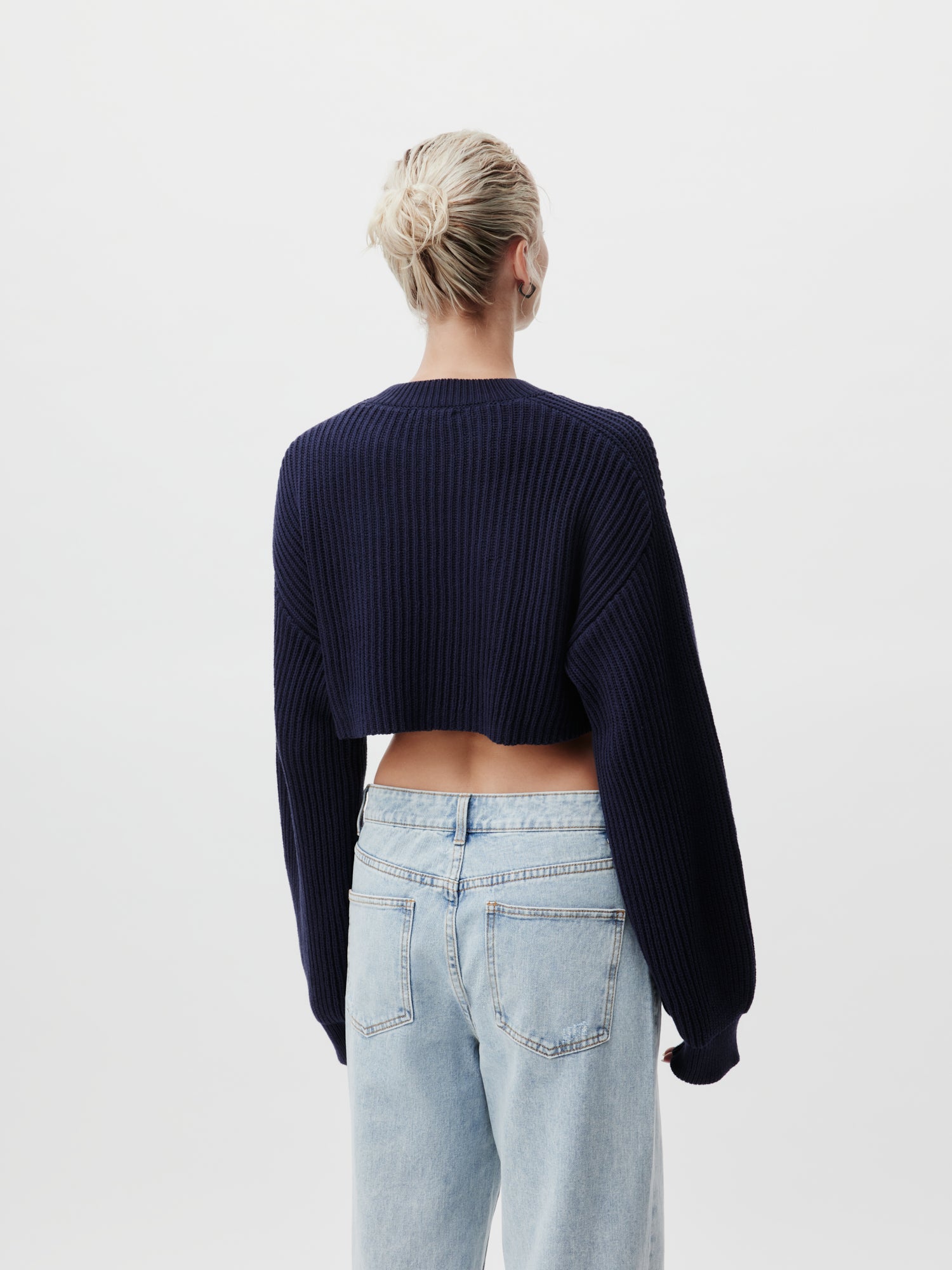 Eine Rückansicht einer Person mit blondem Haar zu einem Dutt, die den Thassia Jumper von LeGer by Lena Gercke trägt, einen marineblauen kurzen Pullover mit langen Ärmeln, kombiniert mit hellblauen Jeans. Der Hintergrund ist schlicht weiß.