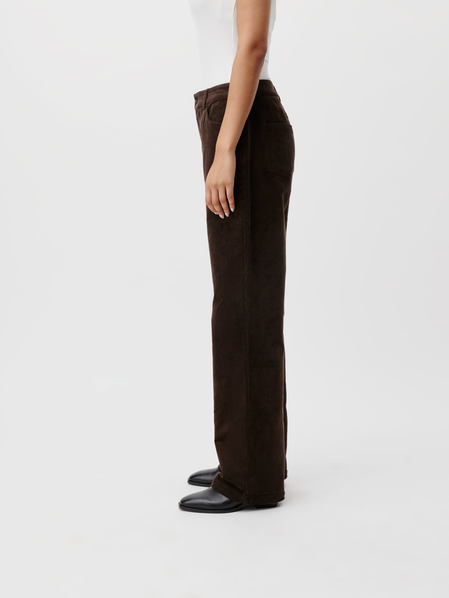 Eine Person in einem weißen Oberteil und der Elora Pants von LeGer by Lena Gercke aus braunem Cord steht im Profil vor einem schlichten weißen Hintergrund. Das 1,77 m große Model ergänzt sein Outfit mit schwarzen Schuhen, die seine Seitenansicht betonen.