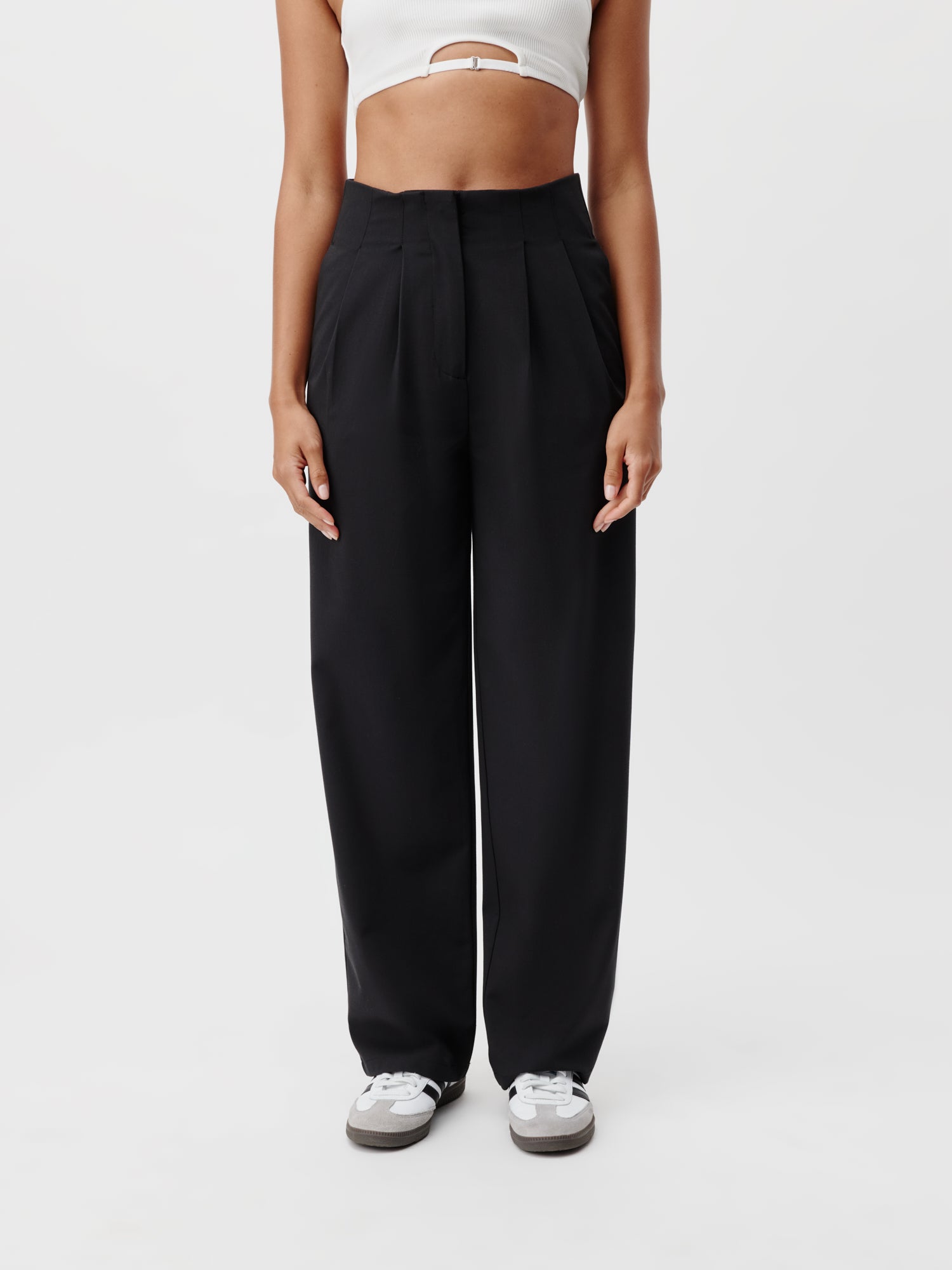 Eine Person trägt die hoch taillierten, schwarzen Marla Pants von LeGer by Lena Gercke, kombiniert mit einem weißen bauchfreien Top und weißen Sneakers mit grauen Sohlen. Die minimalistische Ästhetik von Lang wird durch den schlichten weißen Hintergrund verstärkt.