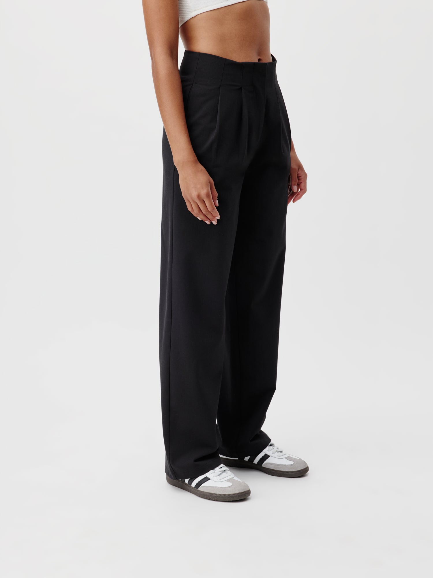 Eine Person, die die Marla Pants von LeGer by Lena Gercke trägt, steht vor einem weißen Hintergrund und präsentiert die mühelose Länge der schwarzen Hose mit weitem Bein. Sie vervollständigen den Look mit schwarzen und weißen Sneakers, die sowohl Passform als auch Stil betonen, und kombinieren sie mit einem weißen bauchfreien Top.