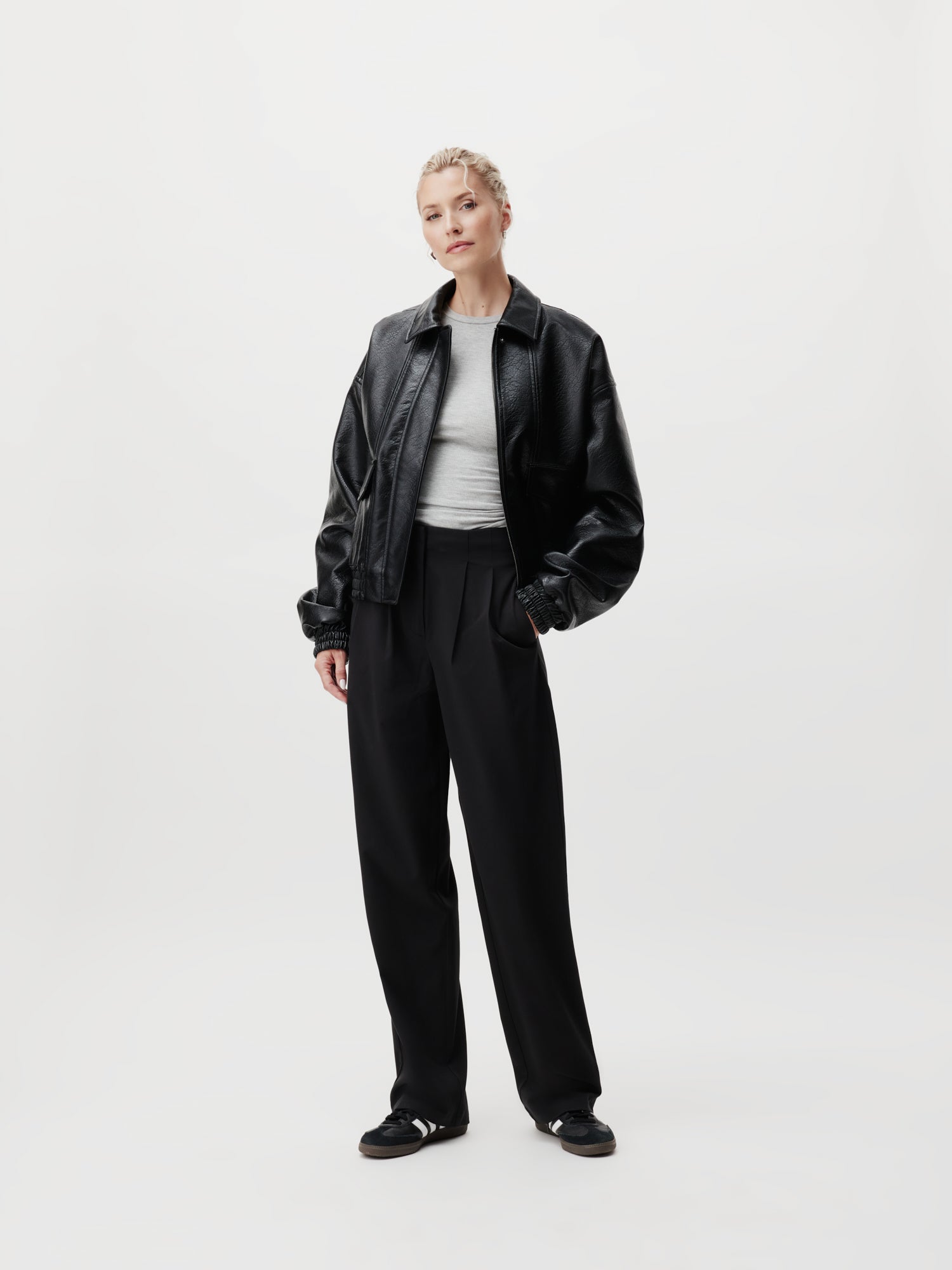 Eine Person strahlt Selbstbewusstsein aus in einer auffälligen schwarzen Lederjacke, einem blütenweißen Hemd und der eleganten Marla Pants Tall von LeGer by Lena Gercke in Schwarz, komplett mit Maxi-Touch. Ihr kurzes, helles Haar bildet einen schönen Kontrast zu einem schlichten hellen Hintergrund und fängt zeitlose Eleganz in Größe 38 perfekt ein.