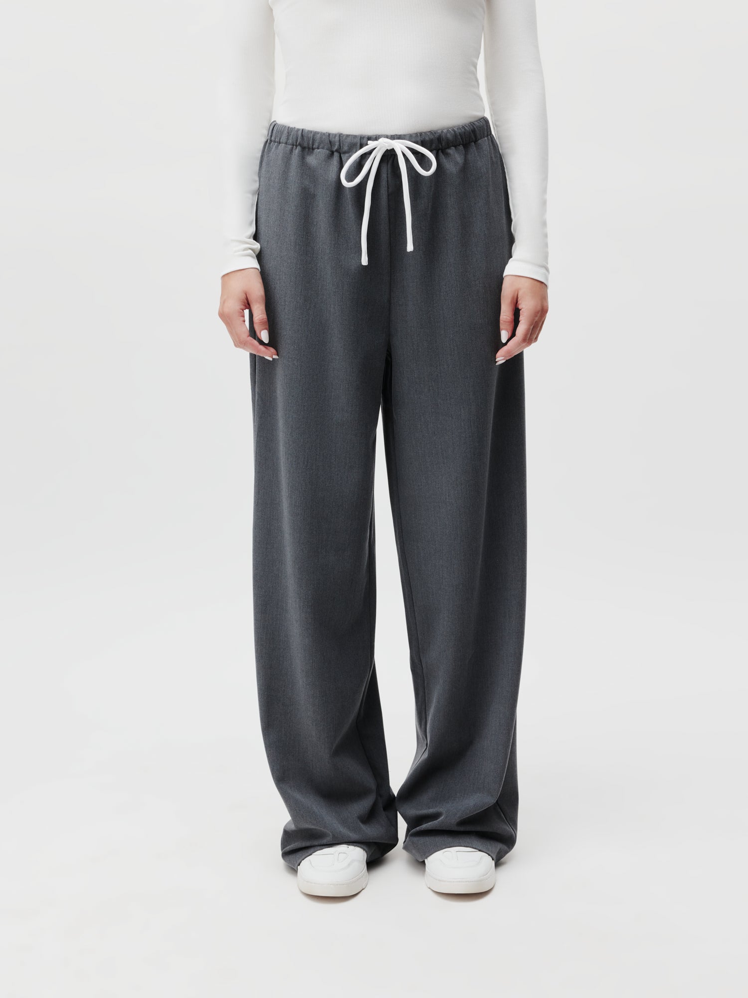 Ein Model präsentiert sich in einem Ensemble bestehend aus einem langärmeligen weißen Shirt und Kim Pants von LeGer by Lena Gercke in Grau mit Kordelzug, Größe 38, vor einem unifarbenen Hintergrund. Weiße Sneakers komplettieren das Outfit und die Arme hängen entspannt an den Seiten.