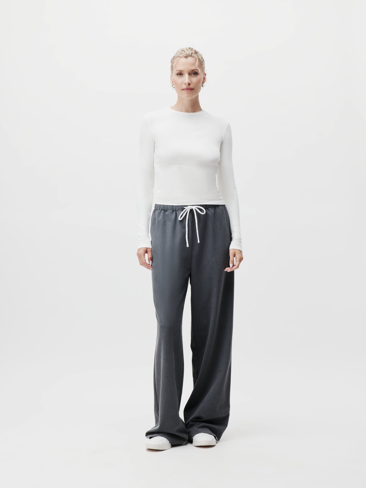 Vor einem schlichten weißen Hintergrund steht ein Model elegant da und präsentiert ein tailliertes weißes Langarmshirt, kombiniert mit der locker sitzenden Kim Pants von LeGer by Lena Gercke in Dunkelgrau mit weißem Kordelzug. Stilvolle weiße Schuhe runden den Look ab. Das Model trägt Größe 38, was die perfekte Passform und den Fluss des Outfits unterstreicht.